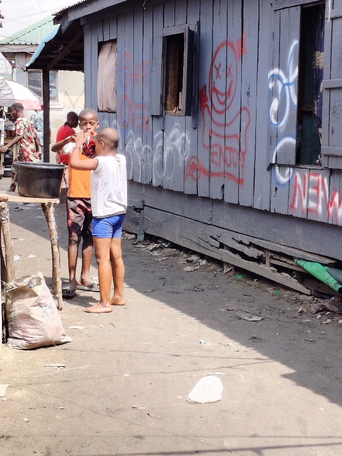 The scene depicts an outdoor urban environment in Oworonshoki, Nigeria, under direct sunlight. In the foreground, two young males interact on an unpaved dirt path. The taller male on the left wears an orange t-shirt and patterned shorts, with dark sandals, and holds an unidentified object to his face while facing the other boy. The shorter male on the right, with a partially bald head, wears a white t-shirt and blue shorts, and is barefoot, with one arm raised. A black plastic basin rests on a makeshift wooden stand to their left. A large, light-colored sack is on the ground near the stand.
To the right, a single-story building constructed from weathered, light blue/grey horizontal wooden planks occupies the midground. The building features two open windows with dark interiors and wooden shutters. Extensive graffiti covers the wall, including a red spray-painted figure with 'X' eyes and a wide smile, accompanied by the word "ENJOY" below it. White and blue spray-painted text and symbols are also visible, including "NEW" in red and white. The lower section of the building shows deteriorated wooden planks. The dirt ground is strewn with minor debris, including a white plastic item in the foreground and green material near the building's base.
In the background, partially obscured figures, including one in a patterned shirt, are visible near a structure with a blue corrugated metal roof and a white and red striped canopy or umbrella.