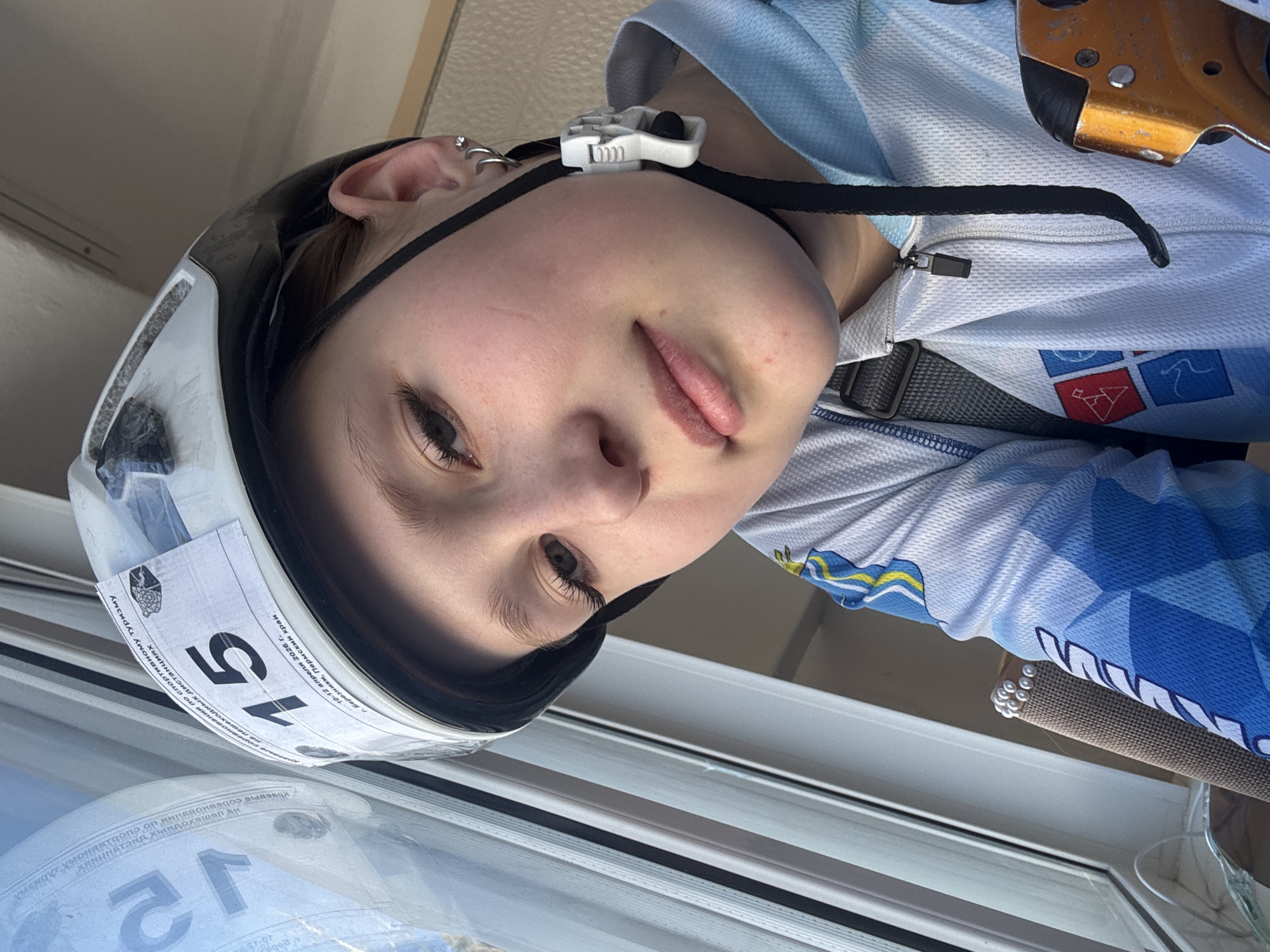 The image shows a person wearing a helmet and a blue and white athletic shirt with patches and an orange device attached. The helmet has a sticker with the number "15" and Cyrillic text identifying the event as a sport tourism competition for walking distances in Berezniki, Perm region, Russia, held on April 10-12, 2026. The person is positioned near a window with blinds.
