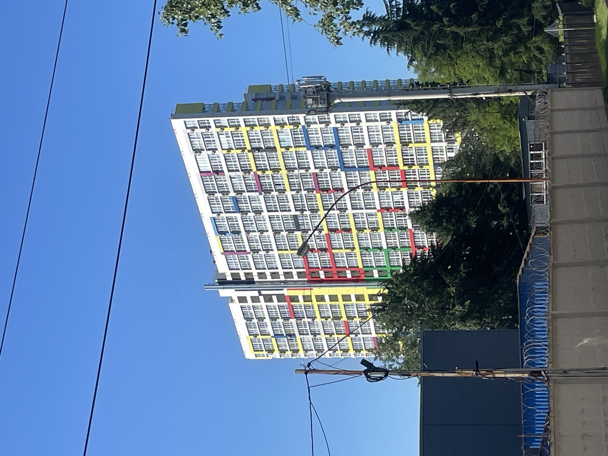 The image, captured in Sochi, Russia, displays a tall, modern apartment building under a clear, bright blue sky on a sunny day. The multi-story structure features a white facade adorned with a distinctive, colorful grid pattern of horizontal and vertical bands in yellow, red, blue, green, and purple, highlighting its numerous windows. Many of these windows have visible air conditioning units.
Green trees with lush foliage are present on both sides of the building, partially obscuring it, with some branches extending into the upper frame. In the mid-ground on the right, a robust fence, likely concrete or masonry, is topped with coils of barbed or concertina wire, indicating a secured boundary. A darker, rectangular structure is visible below this fence. Utility lines stretch across the upper left portion of the scene. No specific activities or individuals are depicted, and no text is visible within the frame. The scene is static, showcasing urban architecture against a natural backdrop.