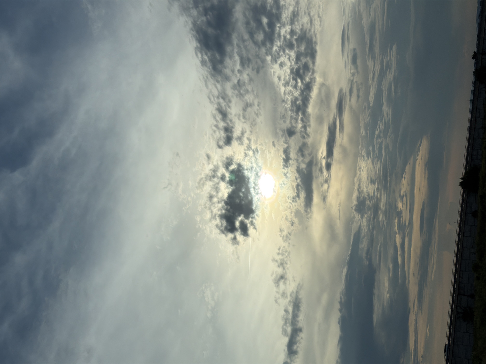 The image depicts a dramatic sky filled with varied cloud formations, ranging from thin, wispy streaks to dense, dark masses. The sun is partially obscured by clouds, creating a soft, diffused glow with a hint of yellow and green. A faint contrail from an aircraft is visible horizontally across the upper-middle section of the sky. In the lower portion of the image, a concrete barrier with a railing runs horizontally, suggesting an elevated roadway or bridge. A single vehicle is visible on this structure, moving from left to right. The overall scene is set against a backdrop of overcast skies, with the sun's light peeking through, casting a somewhat somber yet beautiful atmosphere. The image was taken in Mramor, Bulgaria.