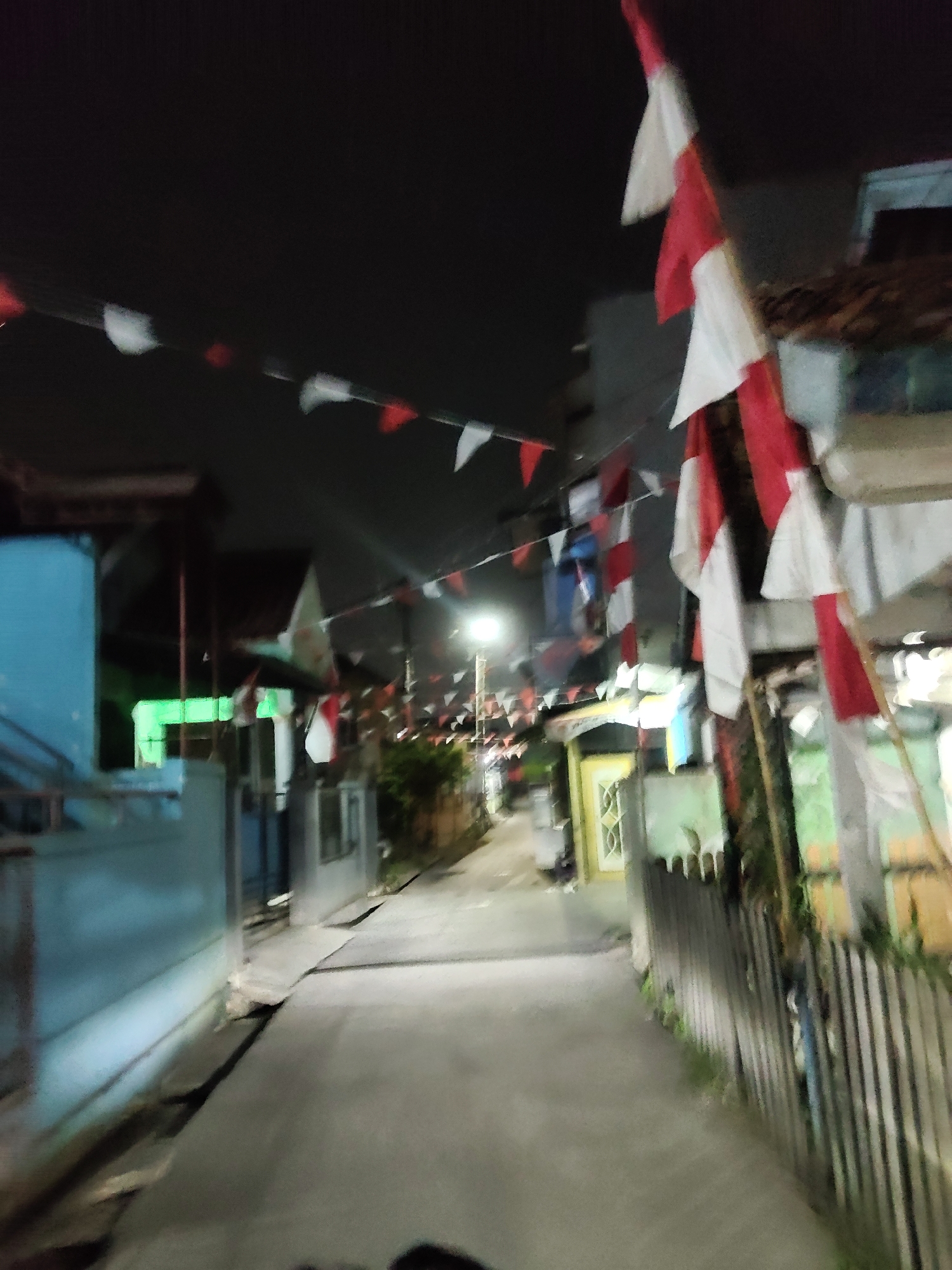 The image captures a narrow, dimly lit street at night in Mekarmukti, Indonesia, decorated with numerous red and white triangular flags strung across it. The flags are in a state of disarray, with some flapping loosely and others appearing tattered. On the left side of the street, a bright blue building is visible, partially illuminated by what appears to be a green light emanating from within. Further down the street, houses with simple facades line both sides. A wooden fence with some overgrown vegetation runs along the right side of the street, partially obscuring the houses behind it. The road surface is paved and appears wet or reflective, suggesting recent rain or a damp environment. The overall atmosphere is one of a residential area during nighttime, likely decorated for a local celebration or event.