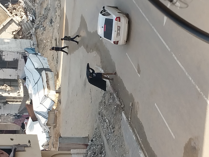 The image depicts a street scene in Khan Yunis, Palestinian Territory, characterized by urban destruction. In the midground, a person wearing a dark shirt, light shorts, and sandals is holding a large, dark, fabric-like object, possibly a tarp or tent section, stretched wide above their head with both hands. To the right of this person, a white sedan, appearing to be a Skoda or similar model, is positioned on the paved road, displaying a license plate partially visible as "569-1XX". Further in the background, two males are walking across the street from left to right; one wears a dark jacket and trousers, while the other wears a dark jacket, dark trousers, and carries a backpack.
The environment features extensive rubble and damaged multi-story concrete buildings in the background. Several makeshift shelters, constructed from white tarpaulins over frames, are visible among the debris behind the street. A partially damaged light-colored building stands on the left edge of the frame. Piles of concrete fragments and dirt line the unpaved shoulder of the road. The paved street itself shows puddles and dirt, with white dashed lines marking lanes. The scene is illuminated by natural daylight.