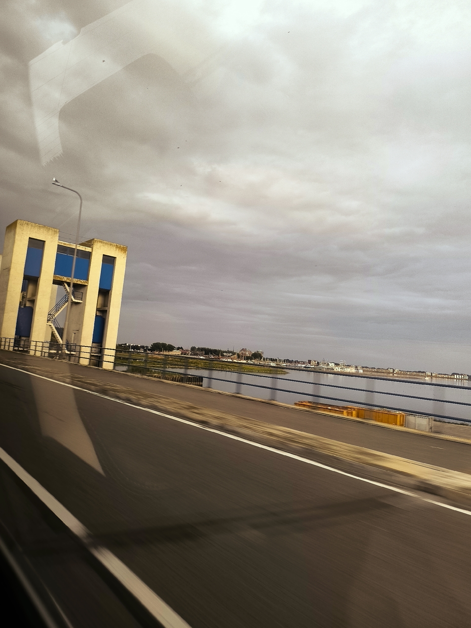 This image captures a view from a moving vehicle on a bridge, likely in Lelystad, Netherlands, given the context. In the foreground, a blurred asphalt road with white markings dominates the lower half of the frame, indicating motion. To the left, a tall, somewhat abstract structure with yellow and blue rectangular panels is visible. This building has an external staircase leading up to its upper section. Beyond the bridge railing, a calm body of water stretches towards a distant cityscape. The cityscape consists of low-rise buildings and structures, appearing to be a waterfront development with some boats docked. The sky above is overcast with heavy, gray clouds, suggesting a potentially gloomy or unsettled weather condition. A street lamp is positioned on the bridge near the building. The overall impression is of a contemporary urban or semi-urban environment seen during transit.