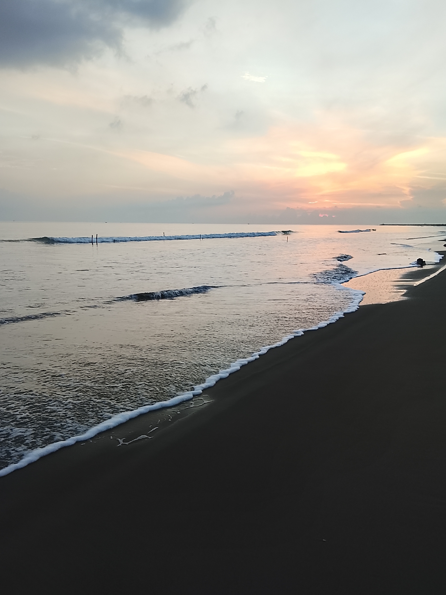 The image depicts a serene beach scene at sunrise. The sky is a soft canvas of pale blues, grays, and warm pinks and oranges, with scattered clouds. The sun is low on the horizon, casting a gentle light across the water and sky. The ocean is calm, with small waves lapping at the shore. The water reflects the colors of the sky, creating a muted, peaceful atmosphere.
The foreground is dominated by dark, wet sand, with a foamy white line of receding waves tracing the edge of the water. In the distance, a few thin, vertical poles are visible in the sea, possibly part of a fishing net or marker. There are also faint silhouettes of what might be distant boats or land structures along the horizon. The overall impression is one of tranquility and the quiet beauty of a new day beginning.