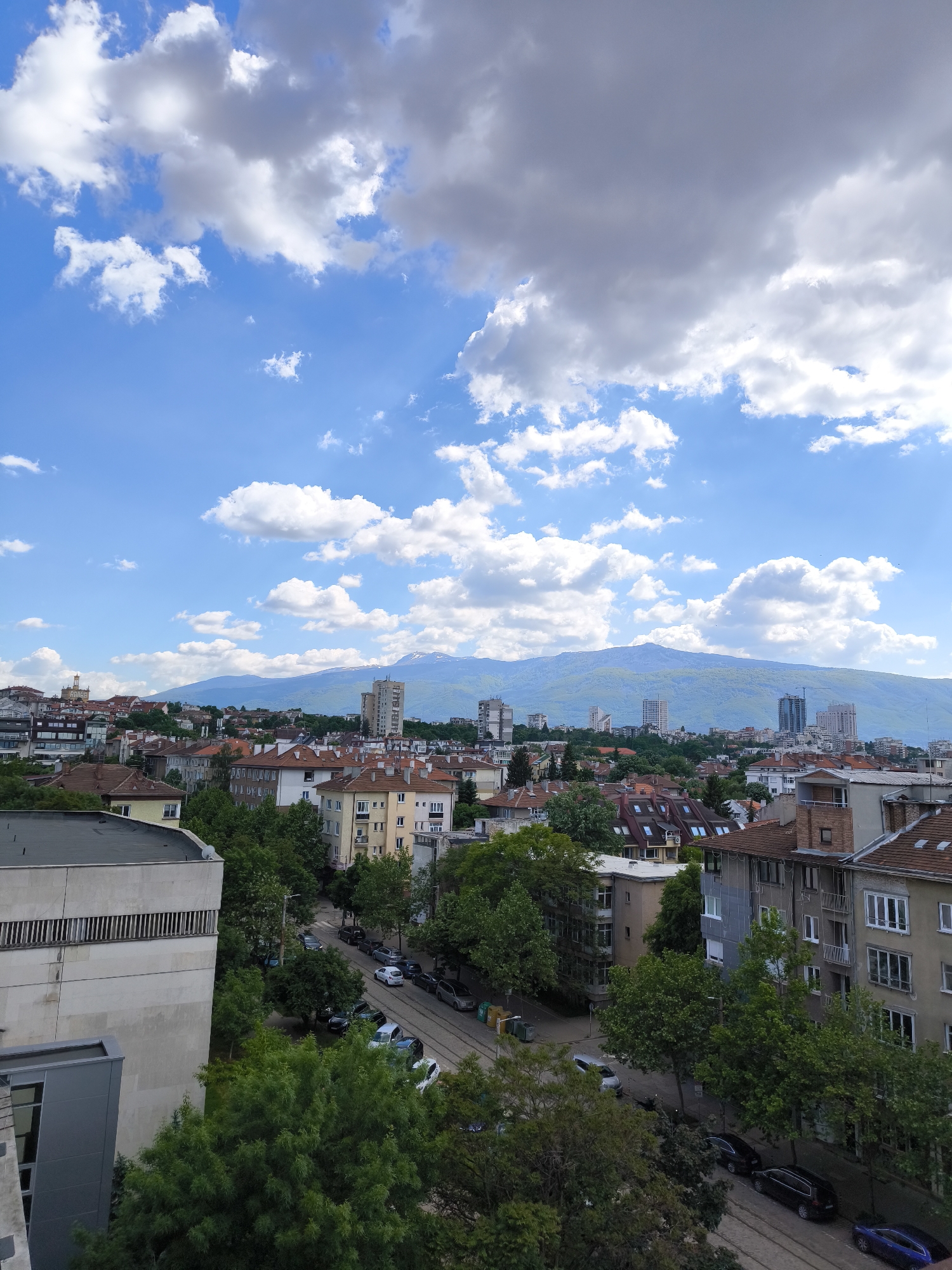 The image shows a cityscape of Sofia, Bulgaria, under a partly cloudy sky. In the background, a mountain range is visible, likely the Vitosha Mountain. The cityscape consists of various buildings, predominantly residential, with red-tiled roofs. In the foreground, there are trees, interspersed with the buildings. Roads with parked cars can be seen between the buildings and trees. The overall impression is an urban scene with a natural backdrop.