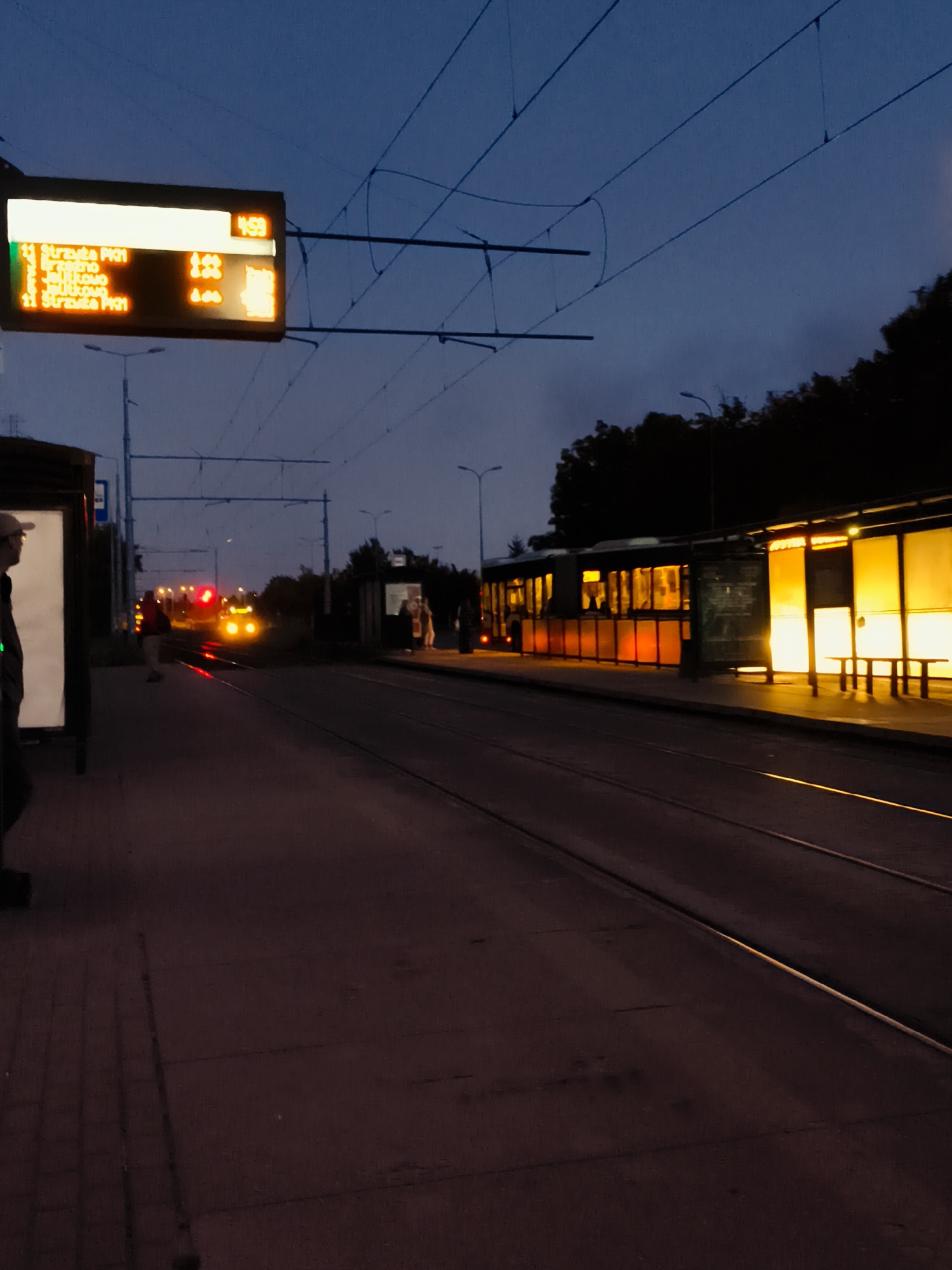 This image depicts a twilight scene at a tram stop in Gdańsk, Poland. The sky is a deep blue, transitioning to lighter hues near the horizon, indicating the late evening or early morning hours. Overhead, electrical wires for the tram system are visible, stretching across the frame.
In the foreground, on the left, a person wearing a cap is partially visible, standing near a large display board. This display board shows tram information, including route numbers (e.g., "11 Strażda PK1"), destinations (e.g., "Brzeźno," "Oliwa," "Jelitkowo"), and departure times (e.g., "4:59," "4:44"). The illuminated text is clearly legible.
The tram stop itself features a shelter with a bright yellow, translucent roof, casting a warm glow on the platform. Benches are visible within the shelter.
The tram tracks run along the right side of the image, leading into the distance. A tram is seen approaching in the middle ground, its headlights and taillights illuminated. The tram appears to be at a stop or approaching one, as its lights are red and yellow.
In the background, trees form a dark silhouette against the fading light. Streetlights are also present, adding to the ambient illumination of the scene. The overall atmosphere is quiet and serene, typical of public transport infrastructure at dusk.