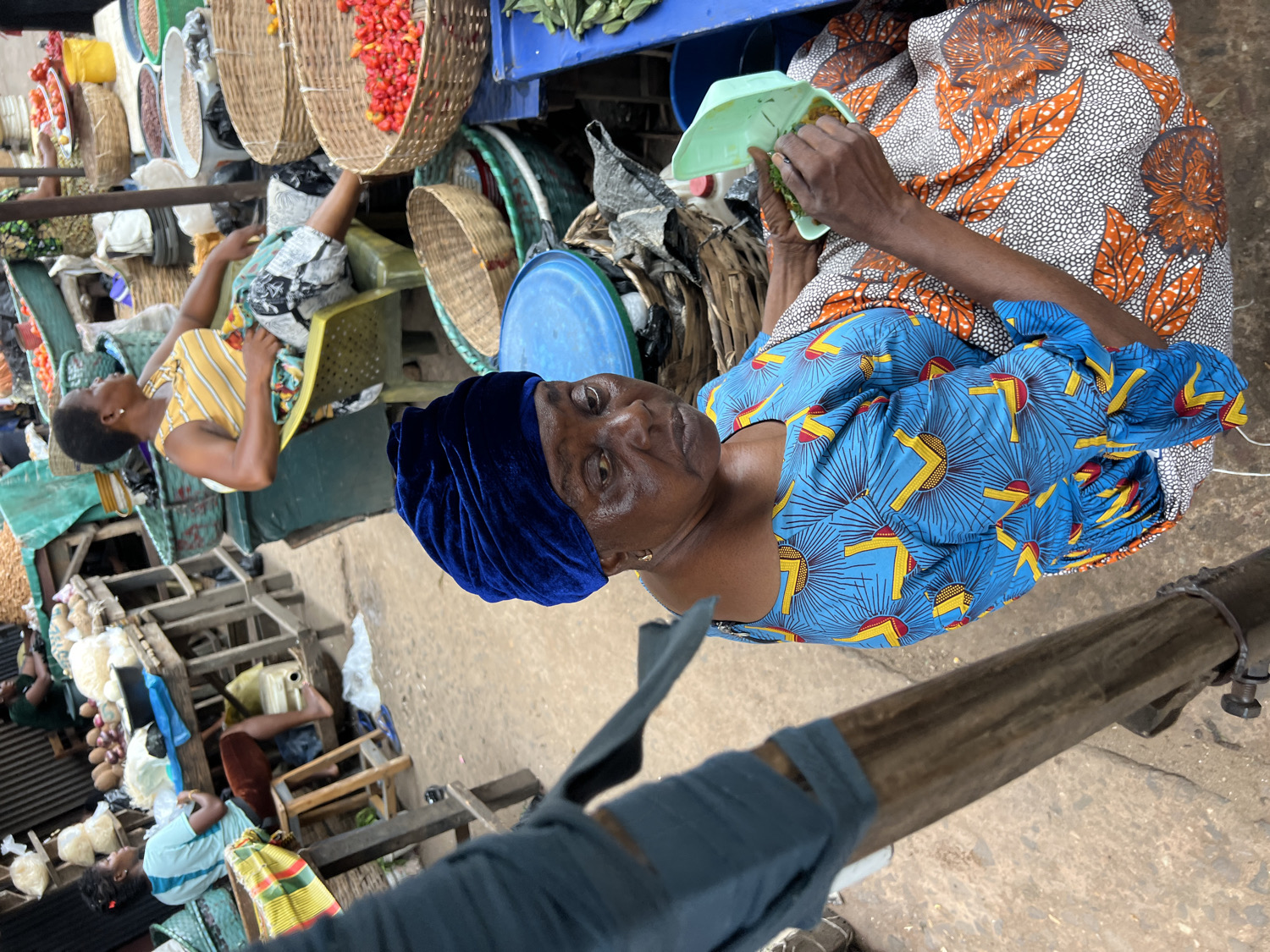An outdoor market scene in Nkpor, Nigeria, captured from a slightly elevated perspective, shows several people amidst market stalls laden with produce. The image is oriented vertically, displaying subjects sideways.
In the immediate foreground, a woman wears a vibrant blue top with yellow and red geometric patterns, and a dark blue velvet head wrap. She looks directly towards the viewer with a neutral expression, holding a light green plastic scoop or small container with some green herbs or vegetables. Her left arm rests on her lap, revealing a patterned skirt or wrapper in orange, white, and black with abstract designs, consistent with West African fabric styles.
Behind her and to the left, another woman in a yellow and white striped top sits relaxed in a green plastic chair, looking to her right. Her stomach appears rounded, possibly indicating pregnancy.
The market stalls are constructed from simple wooden frames. Numerous woven baskets, some hanging and some resting on surfaces, display a variety of fresh produce. Bright red peppers spill from one large hanging basket, while others contain white legumes, dark green vegetables (possibly okra), and other unidentified goods. Blue plastic basins or bowls are also visible on the stalls. In the background, more people are seen among similar wooden structures, some sitting, some attending to their wares. The ground beneath the stalls is a mix of concrete and dirt.
The scene depicts a bustling daytime market, characterized by everyday commerce and the vibrant colours of traditional clothing and fresh produce. No discernible text is visible within the image.