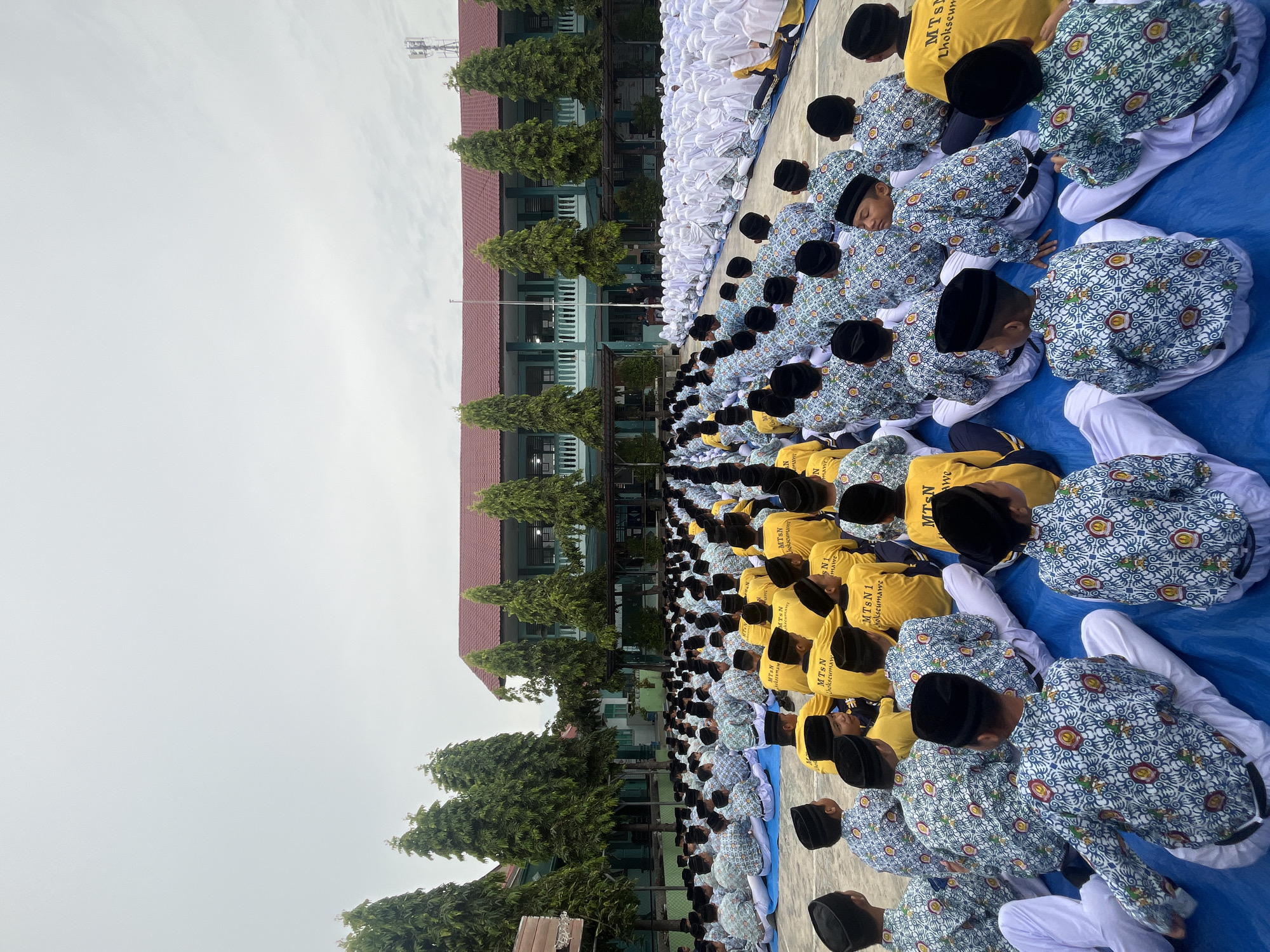 The image captures a wide-angle view of a large group of students, predominantly male, seated on blue tarps outdoors. They appear to be gathered in a schoolyard or assembly area. Many students are wearing black peci hats and uniform shirts, some with a blue and white patterned design, while others wear yellow shirts with text that appears to be "MTsN 1 Lhokseumawe" printed on the back. The students are arranged in rows, facing away from the camera, towards a building in the background. This building has a green facade, multiple windows, a red roof, and a covered walkway. Tall, slender trees are planted in front of the building. The sky is overcast and bright, suggesting a cloudy day. A faint glimpse of a cell tower can be seen on the right side of the image, indicating an urban or semi-urban setting. The presence of "Lhokseumawe" on the uniforms strongly suggests the location is the city of Lhokseumawe in Indonesia. The scene depicts an organized gathering, likely a school event, assembly, or ceremony.