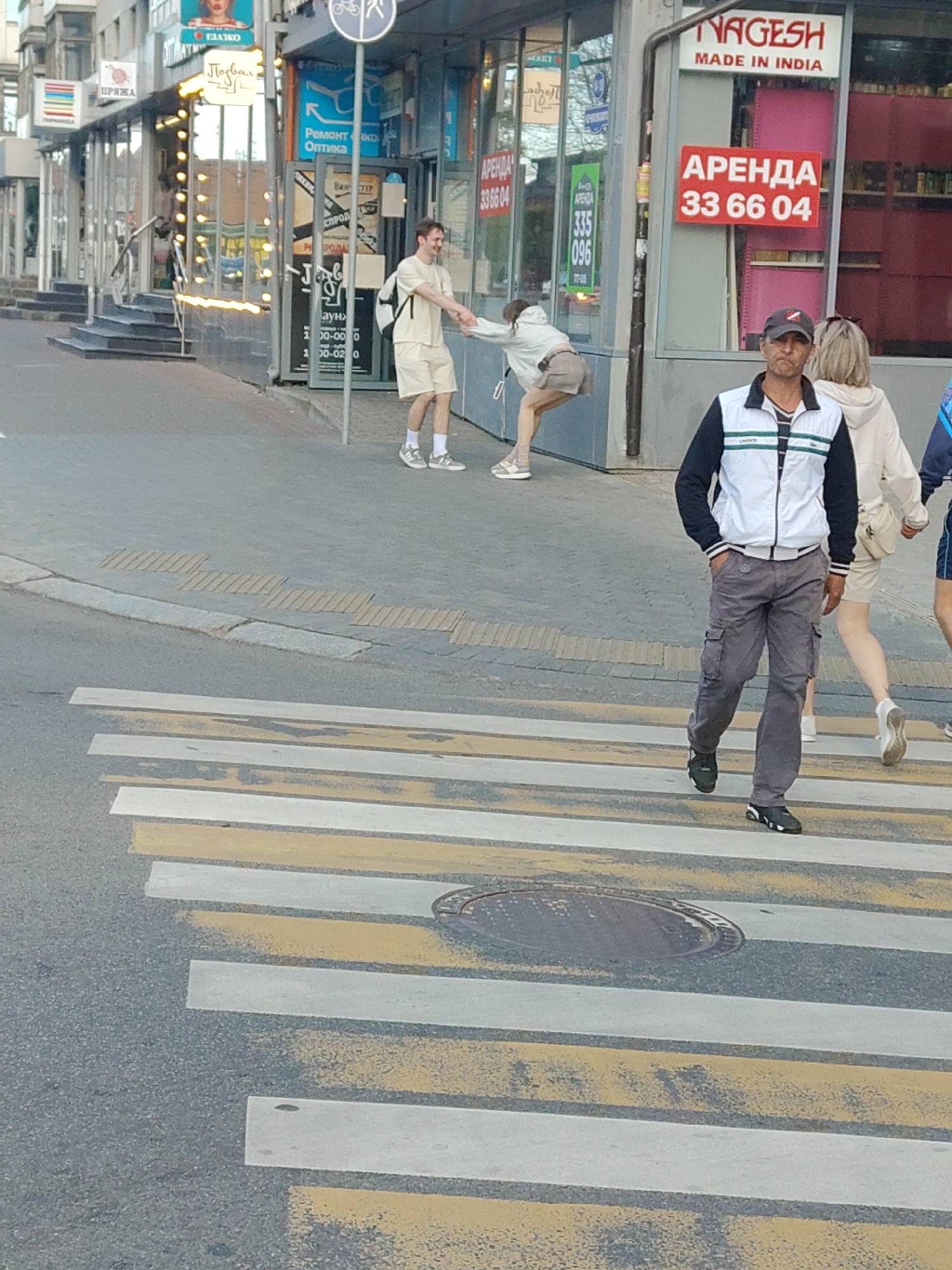 In an urban outdoor setting, a pedestrian crosswalk is prominently featured, marked with alternating yellow and white stripes. To the right of the crosswalk, a man in a white vest with black and green accents and grey cargo pants walks across, his hands in his pockets. He wears a dark baseball cap and dark athletic shoes. Behind him, a woman with blonde hair, wearing a light-colored hooded jacket and white shoes, also crosses the street. Further in the background, two individuals are engaged in an interaction near the entrance of a building with storefronts. One person, wearing a light-colored t-shirt and shorts with a backpack, appears to be handing something to another individual. The building's facade displays various signs, including one with "NAGESH MADE IN INDIA" and another with "АРЕНДА 33 66 04" in red lettering. The environment suggests a commercial area with pedestrian traffic.