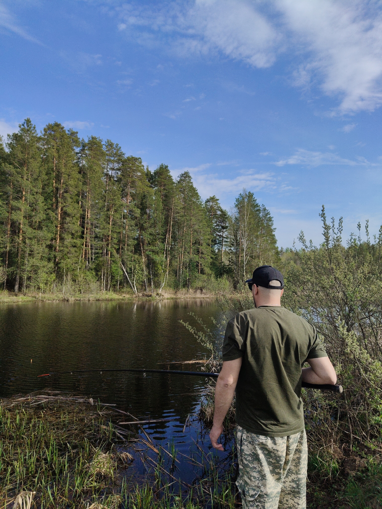 A medium shot depicts a man fishing on a lake. He wears a dark baseball cap, a dark green t-shirt, and camouflage pants. The man stands at the edge of the lake with a fishing rod. The lake is surrounded by a forest of tall trees. The sky is blue with scattered clouds. The location is Unknown, Unknown.