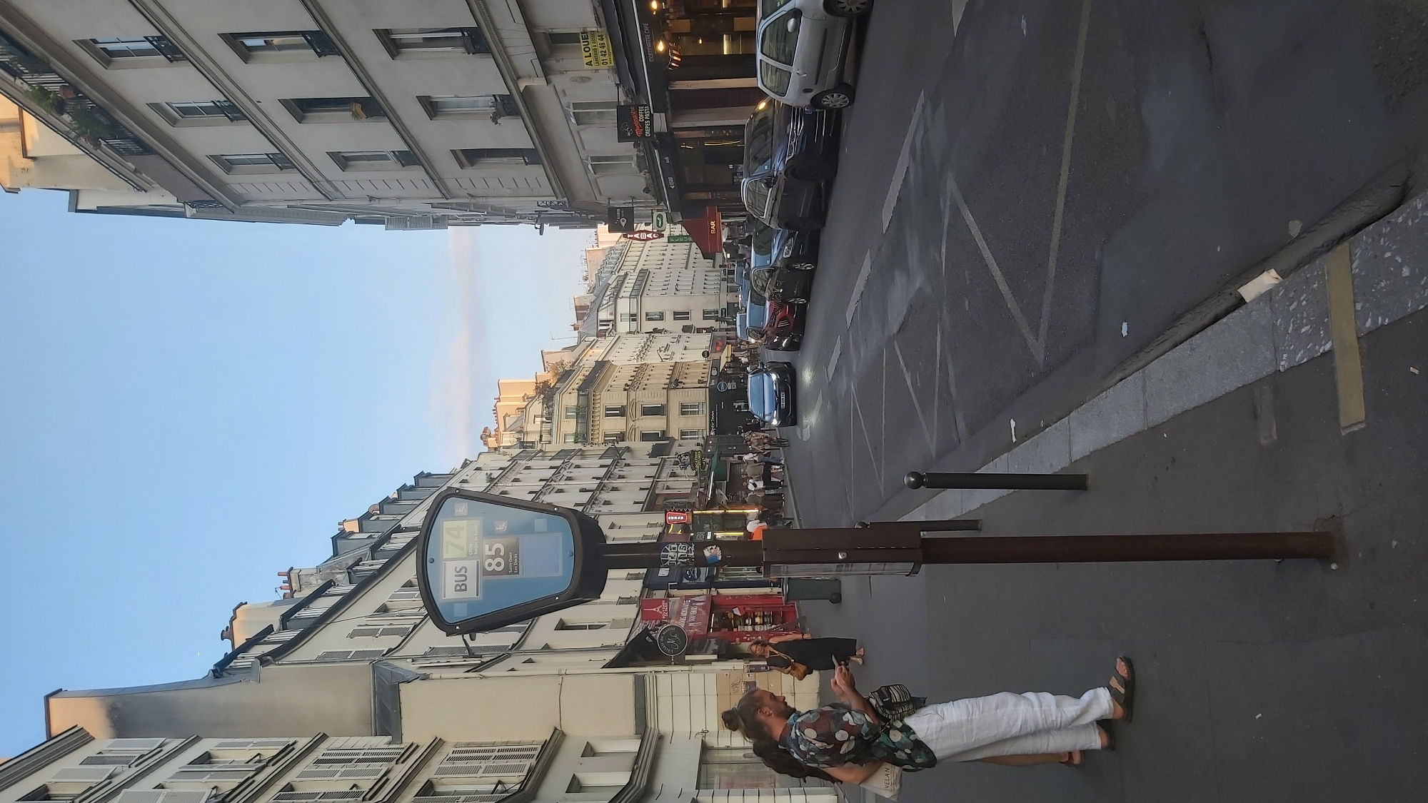 The image depicts a street scene in Paris, France, likely during the late afternoon or early evening. The perspective is from a street-level viewpoint, looking down a moderately busy street lined with Parisian-style buildings. These buildings are multi-story, with light-colored facades, numerous windows with shutters, and pitched roofs. Some buildings feature awnings over storefronts, indicating commercial activity.
In the foreground, a bus stop pole with a digital display is prominently featured. The display shows "BUS 74" and "85 Saint-Ouen Les Docks". Two individuals are standing on the sidewalk near the bus stop. One person, seen from the waist down, is wearing white pants and sandals and is looking at their phone. Another person is partially visible behind them.
The street itself is paved and appears to be wet in places, suggesting recent rain or cleaning. Several cars are parked and in motion on the street. Traffic lights and street signs are visible. Further down the street, more people are walking on the sidewalks and shops are open. The sky above is a clear, light blue with a hint of dusk. The overall atmosphere is one of urban life in Paris.