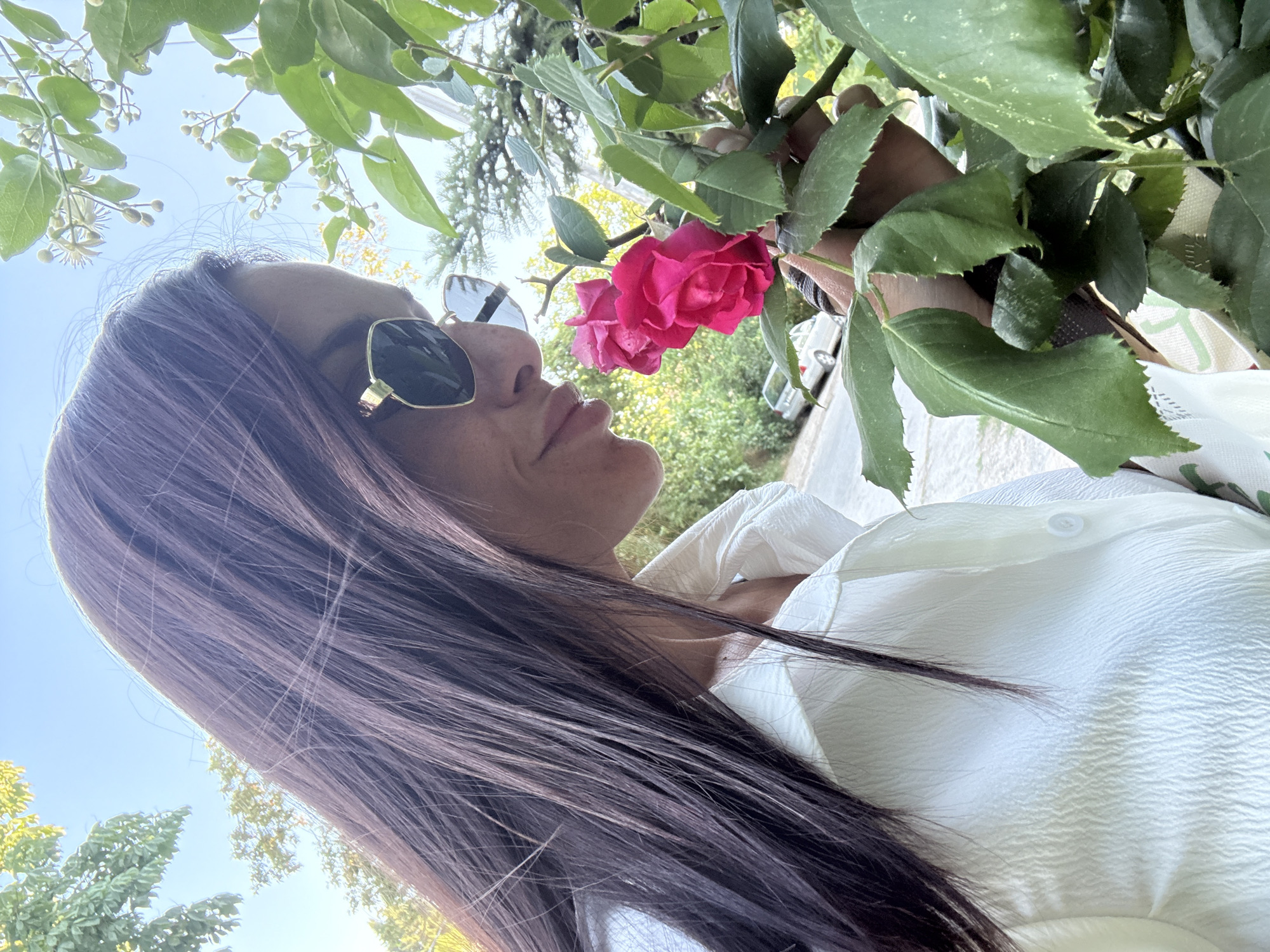 The image shows a close-up, low-angle shot of a woman smelling a pink rose. She is wearing sunglasses with gold frames and a white, textured shirt. Her dark, long hair falls across her shoulder and frames her face. The scene appears to be outdoors, with lush green foliage and branches from a tree overhead, suggesting a garden or park setting. The sky is a clear blue, indicating it is daytime, likely sunny. The woman's face is partially visible, and her eyes are closed as she inhales the rose's scent, conveying a sense of peacefulness and enjoyment. In the background, hints of other greenery and what appears to be a car suggest an urban or suburban environment. The overall impression is one of a serene moment in nature.
