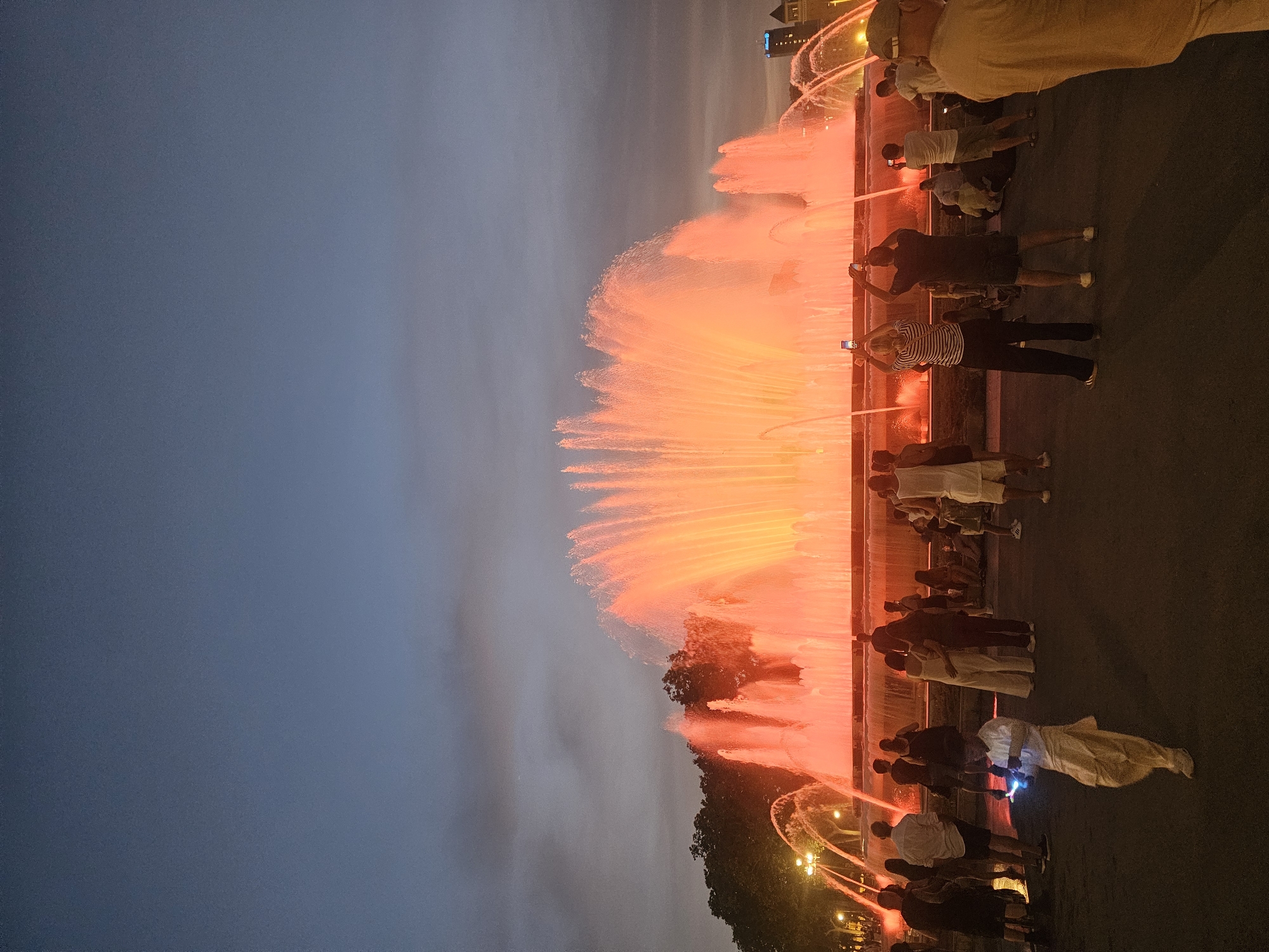 The image depicts the Magic Fountain of Montjuïc in Barcelona, Spain, at dusk. A large, multi-tiered fountain is illuminated with bright orange and red lights, with water jets reaching upwards. A crowd of people is gathered in front of the fountain, many of whom are holding up their phones to take pictures or videos. The sky above is a gradient of dark blue to a lighter hue near the horizon, with some clouds visible. Trees and streetlights can be seen on the left side of the fountain, and buildings are visible in the distance on the right. The ground in the foreground is paved, with people standing and sitting on it. A young boy in a light-colored outfit is walking in the foreground, looking at a device in his hands. The scene captures a public event at a prominent Barcelona landmark.