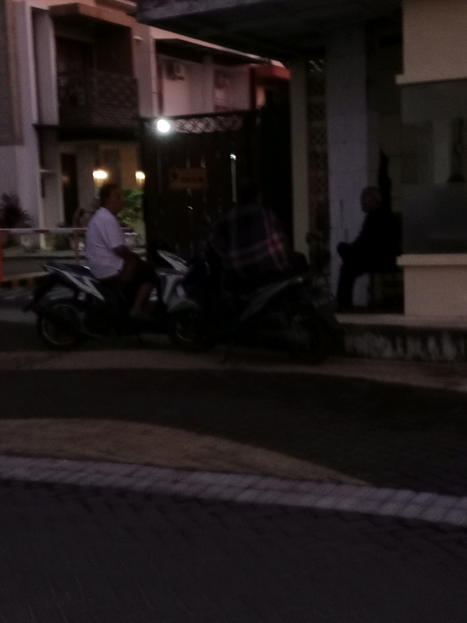 In Waru, Indonesia, at dusk, two men are visible. The first man, wearing a light-colored shirt and dark shorts, is riding a dark-colored scooter. The second man, wearing dark clothing, is seated on a low wall to the right of the first man. Behind them, a multi-story building with a balcony and decorative metalwork is illuminated by lights. The scene is dimly lit, suggesting it is either early evening or late night. The road in the foreground is paved, and there is some vegetation to the left of the building.