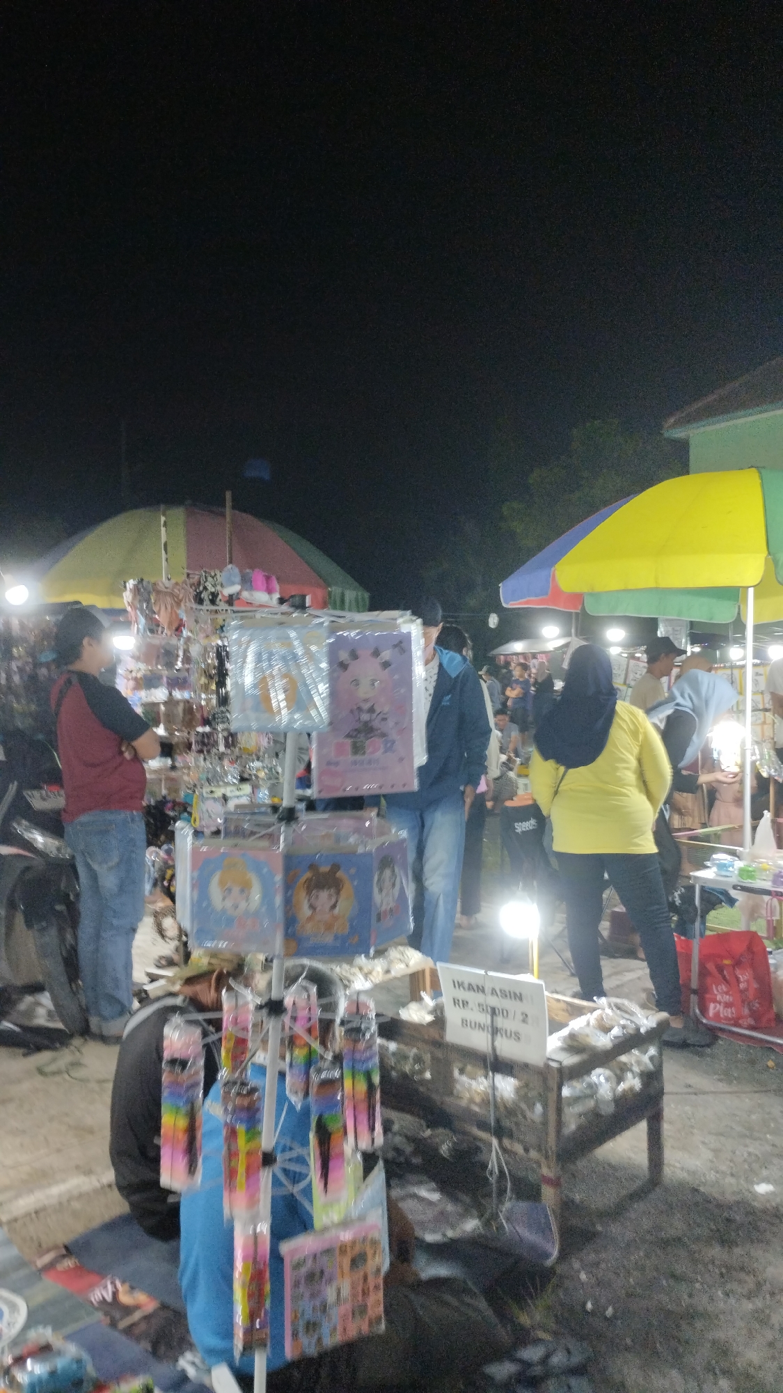 The image captures a nighttime outdoor market scene in Sambungrejo, Indonesia. A stall displays various items, including colorful accessories like hair clips and hair ties packaged in plastic. In front of the stall, a young person wearing a blue shirt and black pants sits on the ground, partially obscured by the display. To the right, a wooden table is laden with packaged dried fish, with a sign above reading "IKAN ASIN RP. 5.000 / 2 BUNGKUS". Several individuals are browsing or working at the market, some standing and others walking, under the illumination of artificial lights. The background features market tents and trees, suggesting a bustling local gathering.