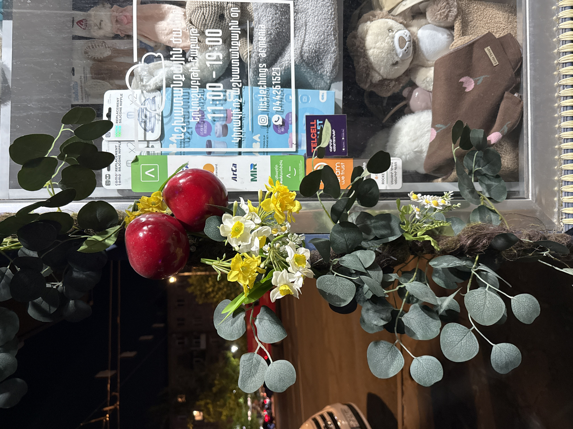 The image shows a storefront in Yerevan, Armenia, at night. The storefront features a window display with various items, including baby products, stuffed animals, and flowers. In the foreground, there is a decorative arrangement of artificial eucalyptus branches and red apples. There is also a yellow and white flower bouquet with daffodils and smaller white flowers.
Behind the flowers and apples, on the glass of the storefront, there are inscriptions in Armenian. One prominent inscription reads: "11:00 - 19:00", indicating the store's operating hours. Below this, it says "littlethings armenia" and a phone number "044261521".
Inside the store, visible through the window, are shelves displaying a variety of items. These include boxes of baby products, a plush lion toy, a knitted blanket with a floral pattern, and other soft toys and baby accessories. The overall impression is that of a shop specializing in children's items. The background shows a street scene at night, with streetlights and building facades dimly visible, suggesting an urban environment.