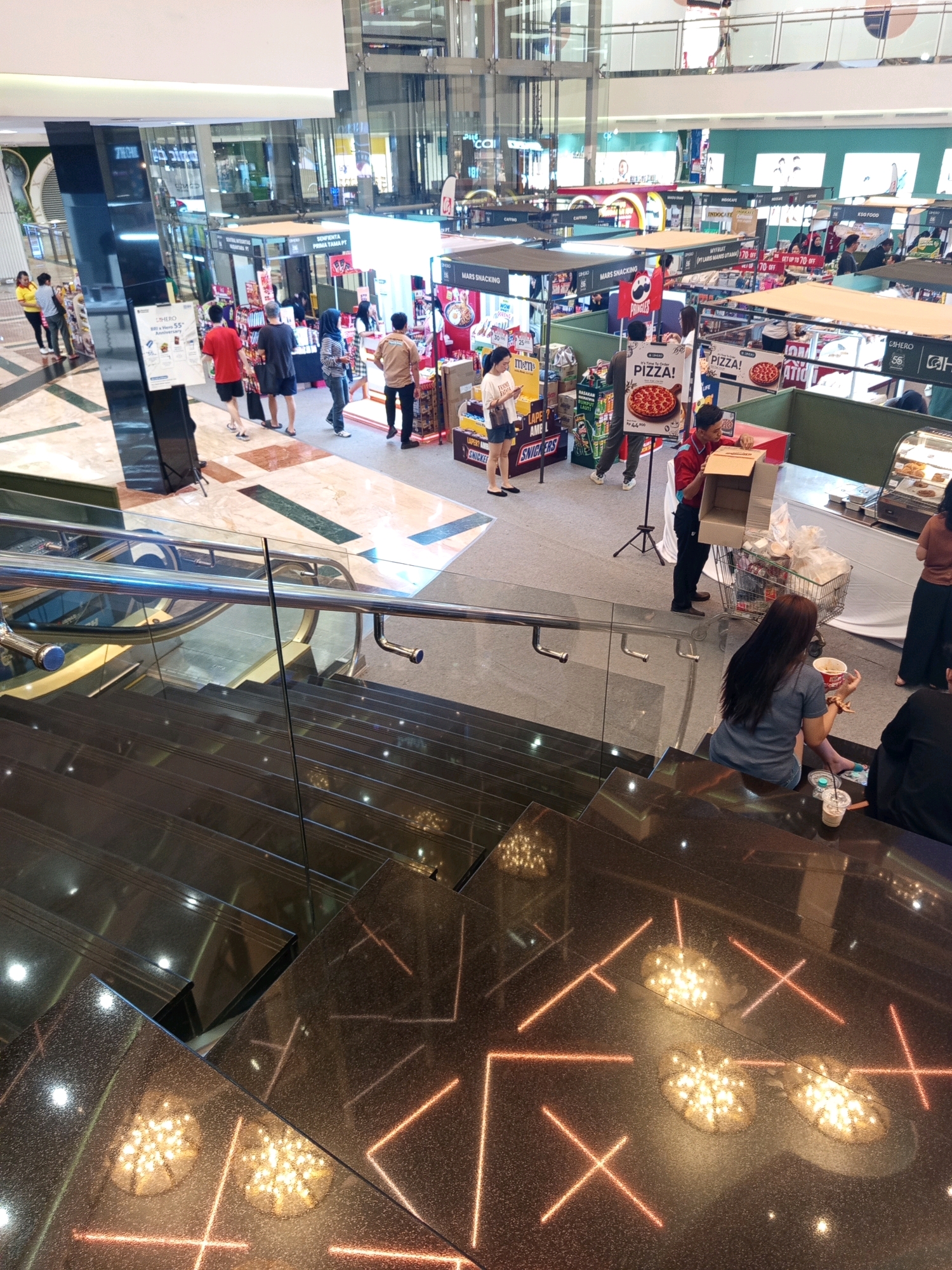 The image captures a bustling scene on the ground floor of Taman Anggrek Mall, located in West Jakarta, Indonesia. The mall's interior features modern architecture with polished floors and escalators. In the foreground, a woman with long dark hair sits on a dark, reflective staircase, holding a cup of food. Next to her, on the stairs, are a disposable cup with a straw and a smaller white cup.
Further into the mall, a variety of stalls and shops are visible. Several people are browsing and shopping. One stall displays a large sign with the word "PIZZA!" and an image of a pizza. A male employee in a red shirt is actively working at this stall, handling a cardboard box and a shopping cart filled with items. Another stall has signage related to "MARS SNACKING." People of various ages and ethnicities are present, engaged in shopping and movement. The environment is well-lit, typical of a large indoor shopping center. Reflections of lights and the surrounding architecture are prominently visible on the dark, glossy surfaces of the stairs and floor.