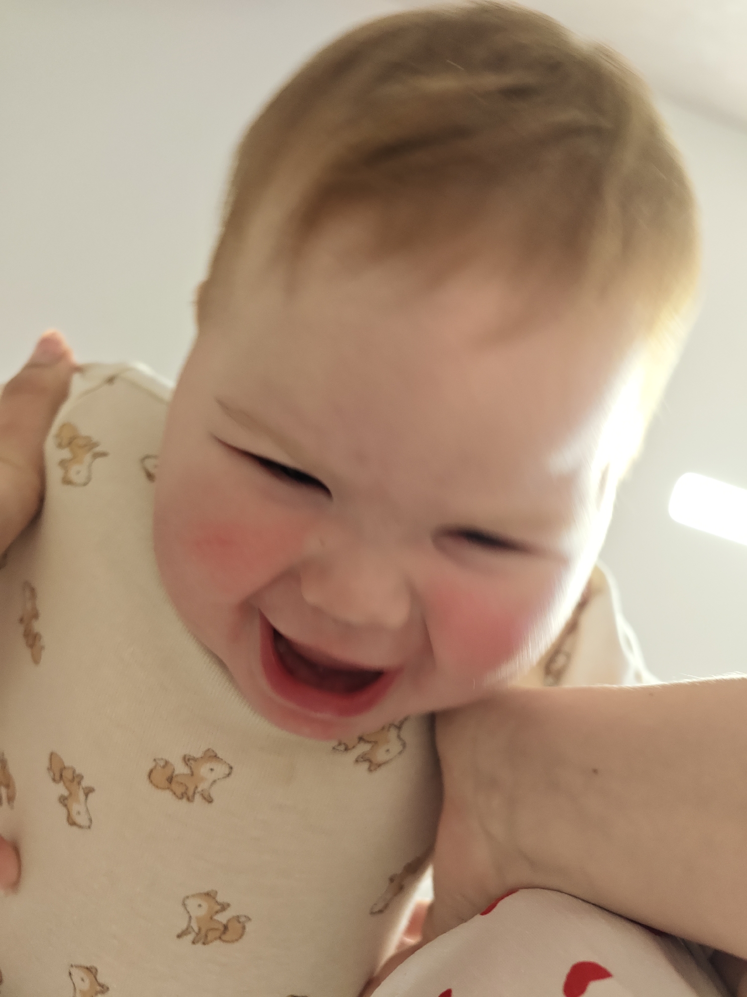 A baby with fair skin and blonde hair is laughing. The baby is being held by someone, as a hand is visible on the left and right sides of the frame. The baby is wearing a light-colored onesie patterned with small fox images. Redness is present on the baby's cheeks. The background is primarily blurred and light. The location is not discernible in the image. The city of Chaikovsky, Russia, cannot be determined from the image.