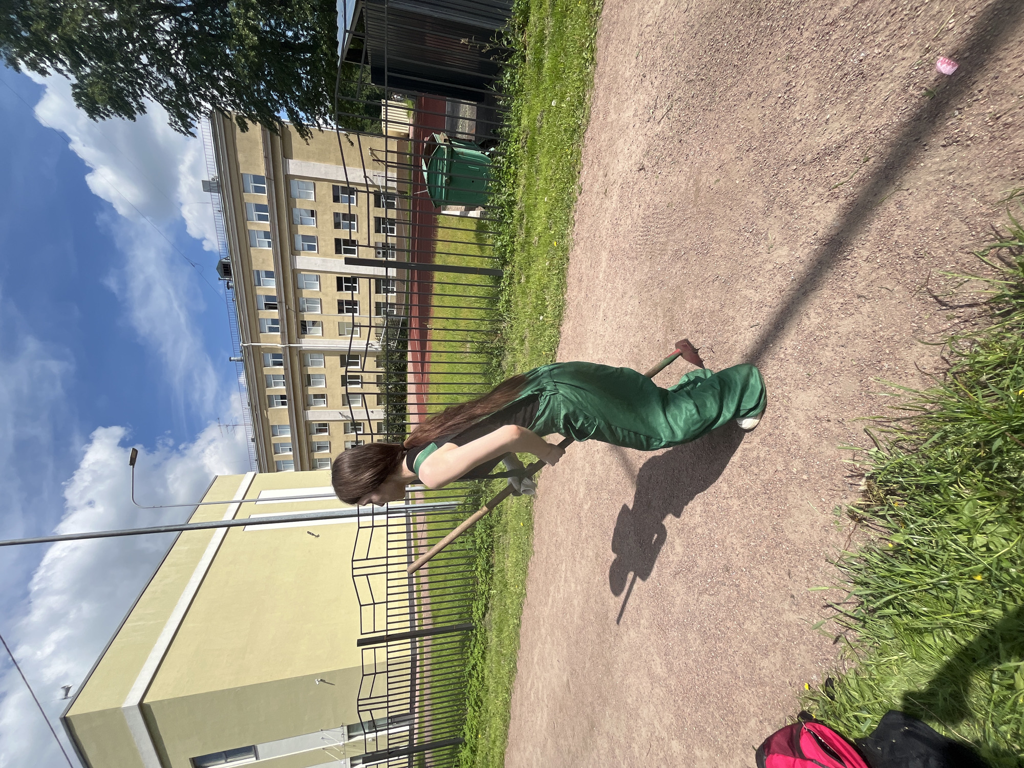 A young woman with long brown hair, wearing green overalls and a dark sleeveless shirt, is captured mid-action on a gravel path in Saint Petersburg, Russia. She is holding two long-handled tools, one in each hand, with the tips of both tools touching the ground. Her stance suggests she is engaged in some form of manual labor or sport, possibly related to grounds maintenance or an outdoor game.
The environment is an outdoor setting, likely a park or school grounds, indicated by the presence of a yellow building in the background, a black metal fence, green grass, and a sports field with a red track. A large, green garbage bin is visible behind the fence, and a dark-colored bag with a pink backpack lies on the grass to the left of the path. The sky is blue with scattered white clouds, and the sun casts a long shadow of the woman and her tools across the gravel. The overall scene depicts a bright, sunny day in a semi-urban, potentially educational or recreational, environment within Saint Petersburg.