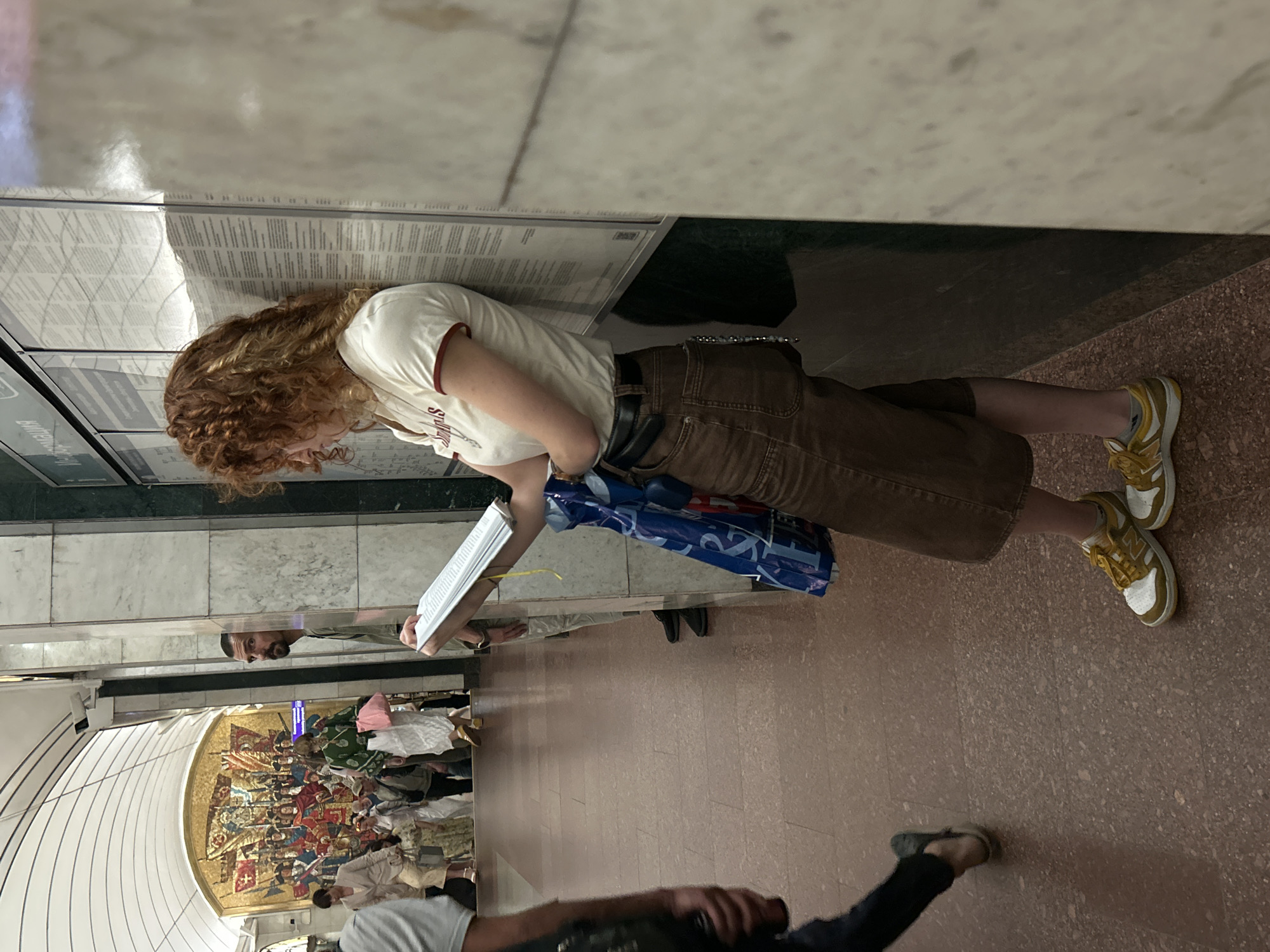 The image shows a young woman with curly red hair standing in what appears to be a metro station in Shushary, Russia. She is leaning against a wall covered with informational posters, holding a book or pamphlet open. She is wearing a white t-shirt with a red trim, brown shorts, and yellow and white sneakers. A blue shopping bag hangs from her left arm. In the background, other commuters are visible on an escalator and walking down a hallway. The station has marble walls and a reddish-brown floor. A large mosaic artwork is visible on the far wall of the escalator area. To the left of the woman, a man with a beard is looking towards the camera.