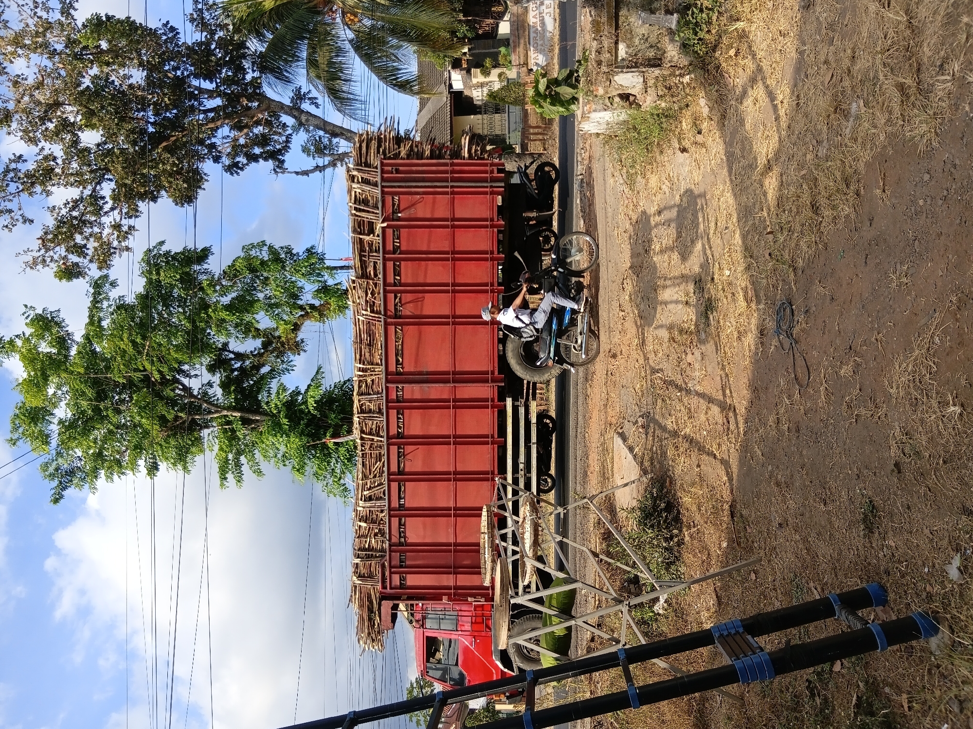 A red truck loaded with wooden poles is parked on a dusty road in Tanggul Wetan, Indonesia. A man on a motorcycle rides past the truck. In the foreground, a ladder and a metal stand with circular objects are placed on the side of the road. Trees and power lines are visible in the background. The sky is blue with scattered clouds.