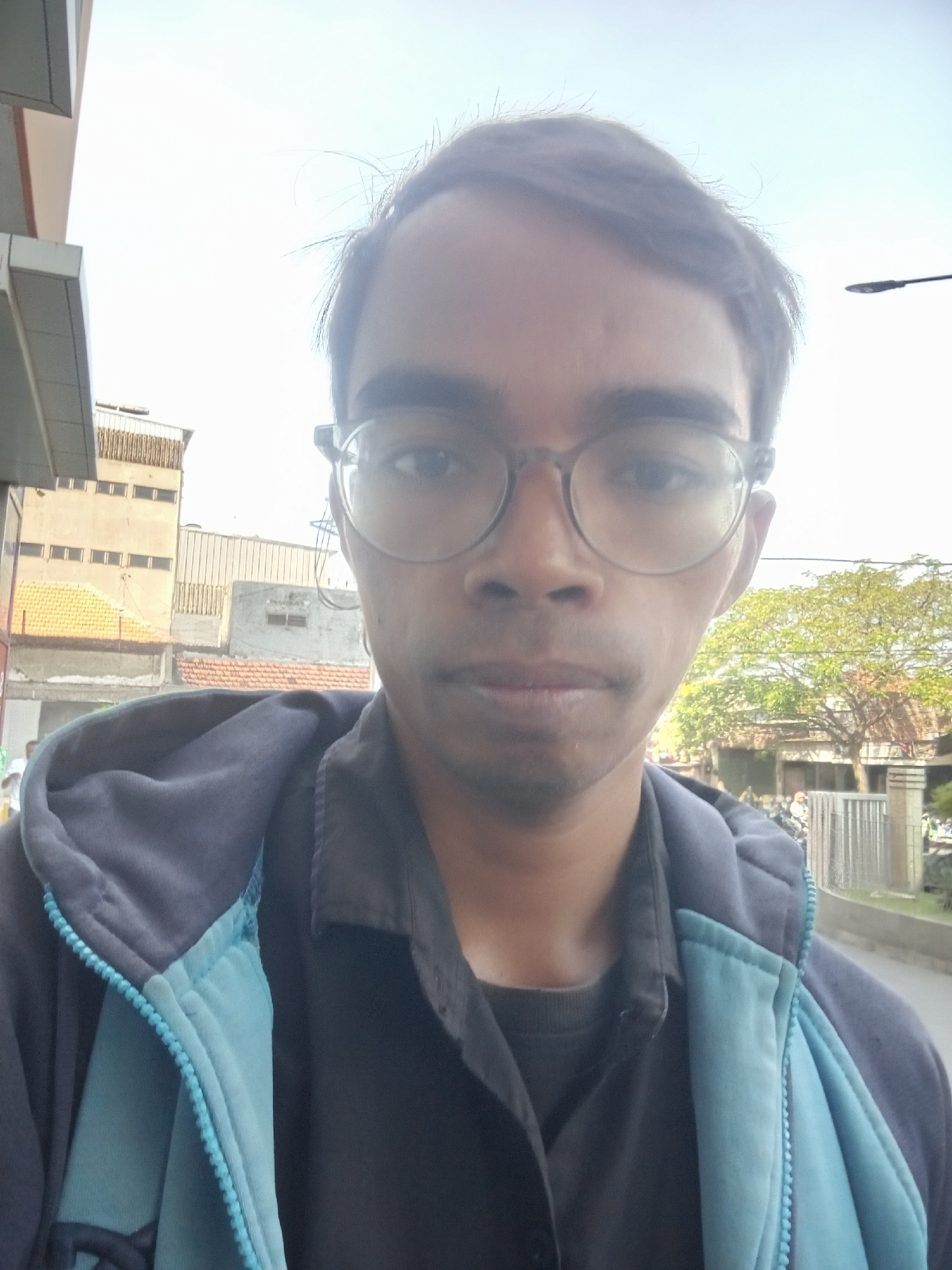 A man with glasses and a black shirt under a blue jacket is looking at the camera. He is outside, in front of buildings and trees. The sky is bright. The location is likely Surabaya, Indonesia, as indicated by the architecture and vegetation. The overall scene is a casual outdoor portrait, possibly taken on a street or sidewalk.
