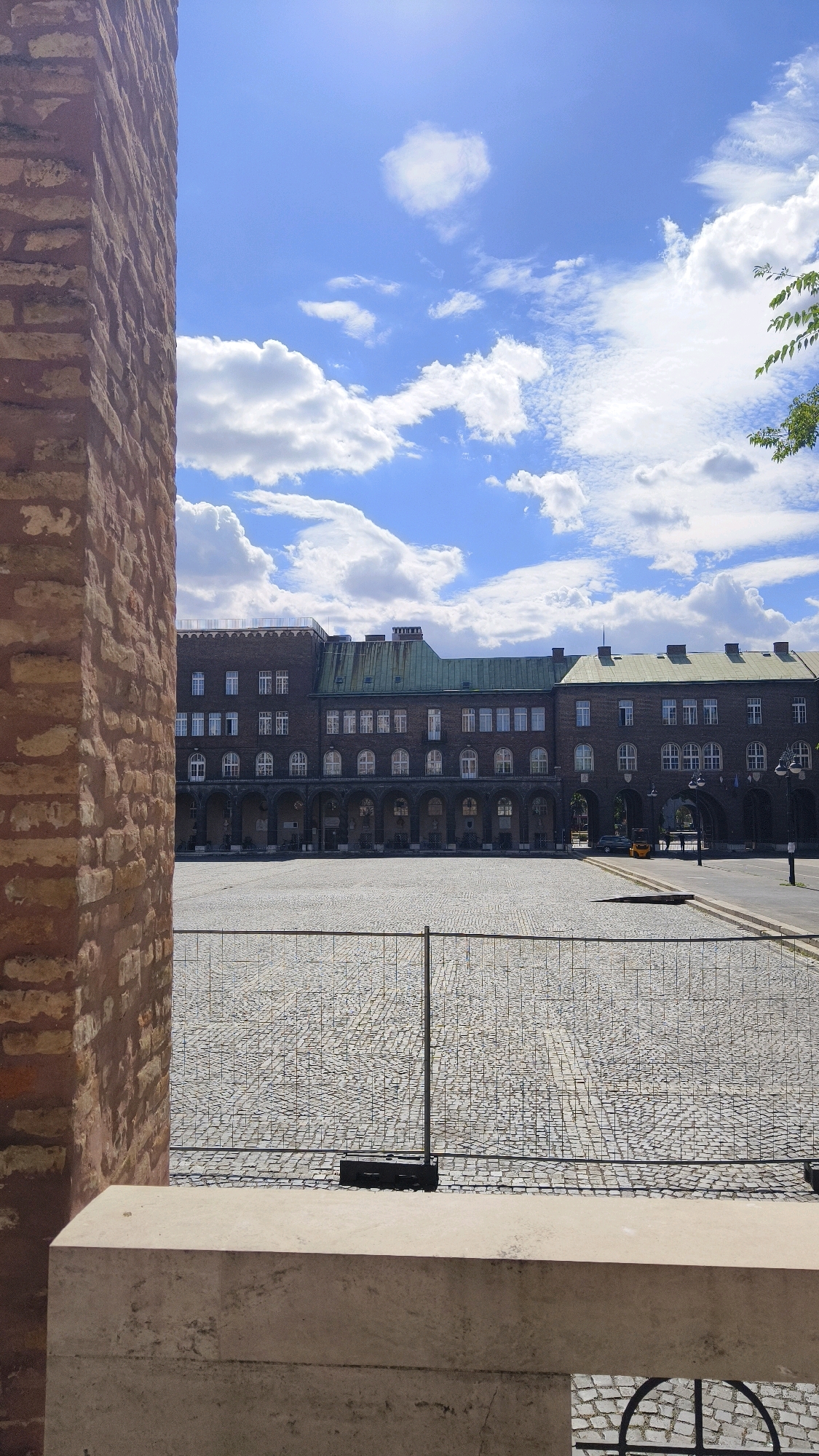 This is a medium shot of the exterior of a large, multi-story building in Szeged, Hungary. The building has a brick facade and a green roof. The front of the building features a series of arches on the ground floor. In front of the building is a large, cobblestone courtyard. A temporary fence is erected in the courtyard. The sky is blue with scattered white clouds. A portion of a brick wall is visible on the left side of the image, and a concrete ledge is in the foreground.