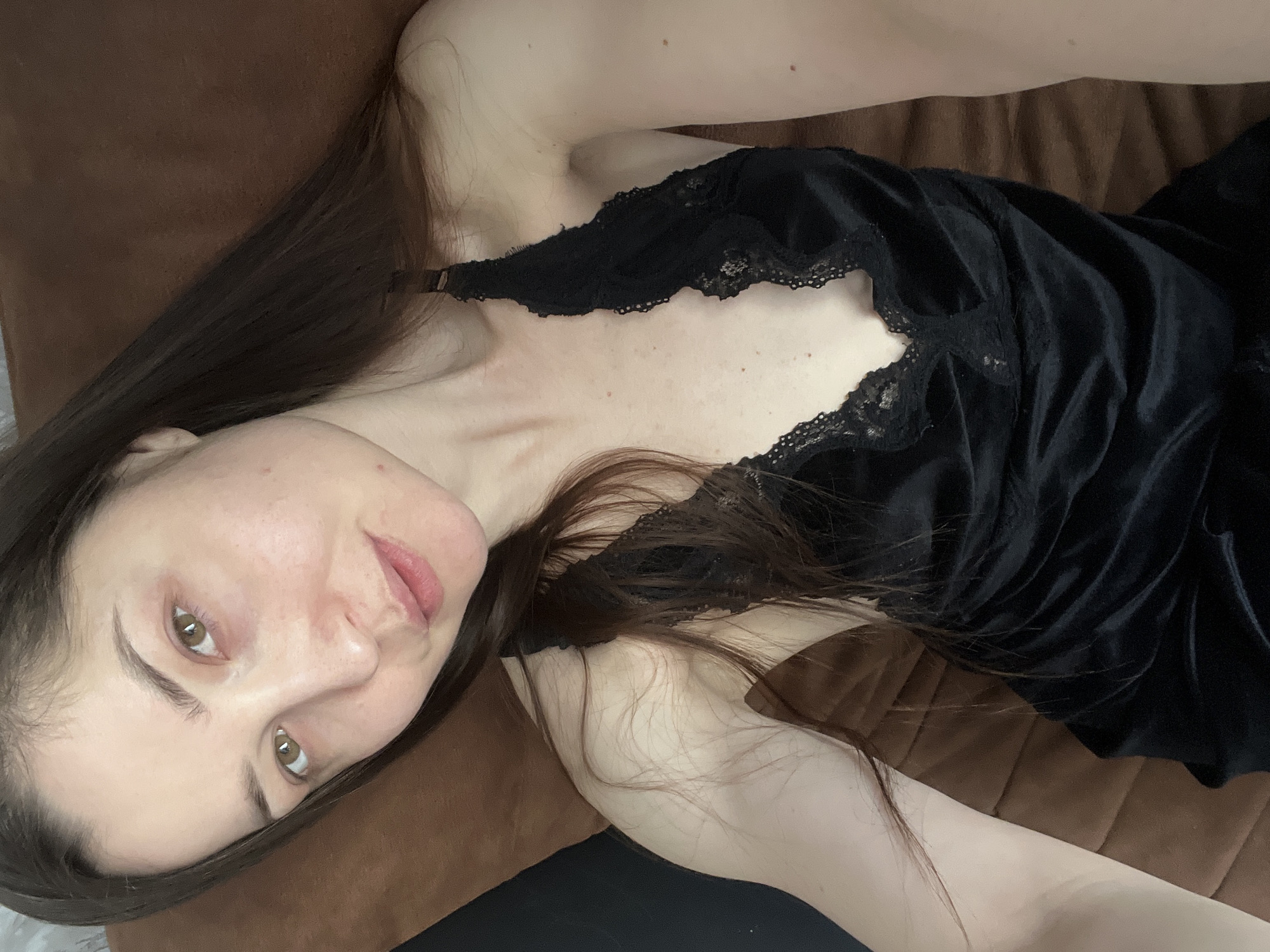 The image features a woman lying down on a brown, upholstered surface, possibly a couch or bed. Her body is positioned diagonally across the frame, with her head in the lower-left corner looking up and towards the camera. She has long, straight dark brown hair that falls around her shoulders and back.
The woman is wearing a black top, which appears to be a camisole or nightgown, made of a soft, possibly velvet or satin-like fabric, with delicate black lace trim along the neckline. Her skin is fair, and some natural blemishes, such as small red spots on her chest and shoulder, are visible. Her facial expression is neutral to slightly contemplative, with a subtle hint of a smile. She has light brown or hazel eyes and naturally defined eyebrows. The lighting is soft and even, highlighting her features. No visible text is present in the image. The scene conveys a relaxed and intimate atmosphere, likely indoors.