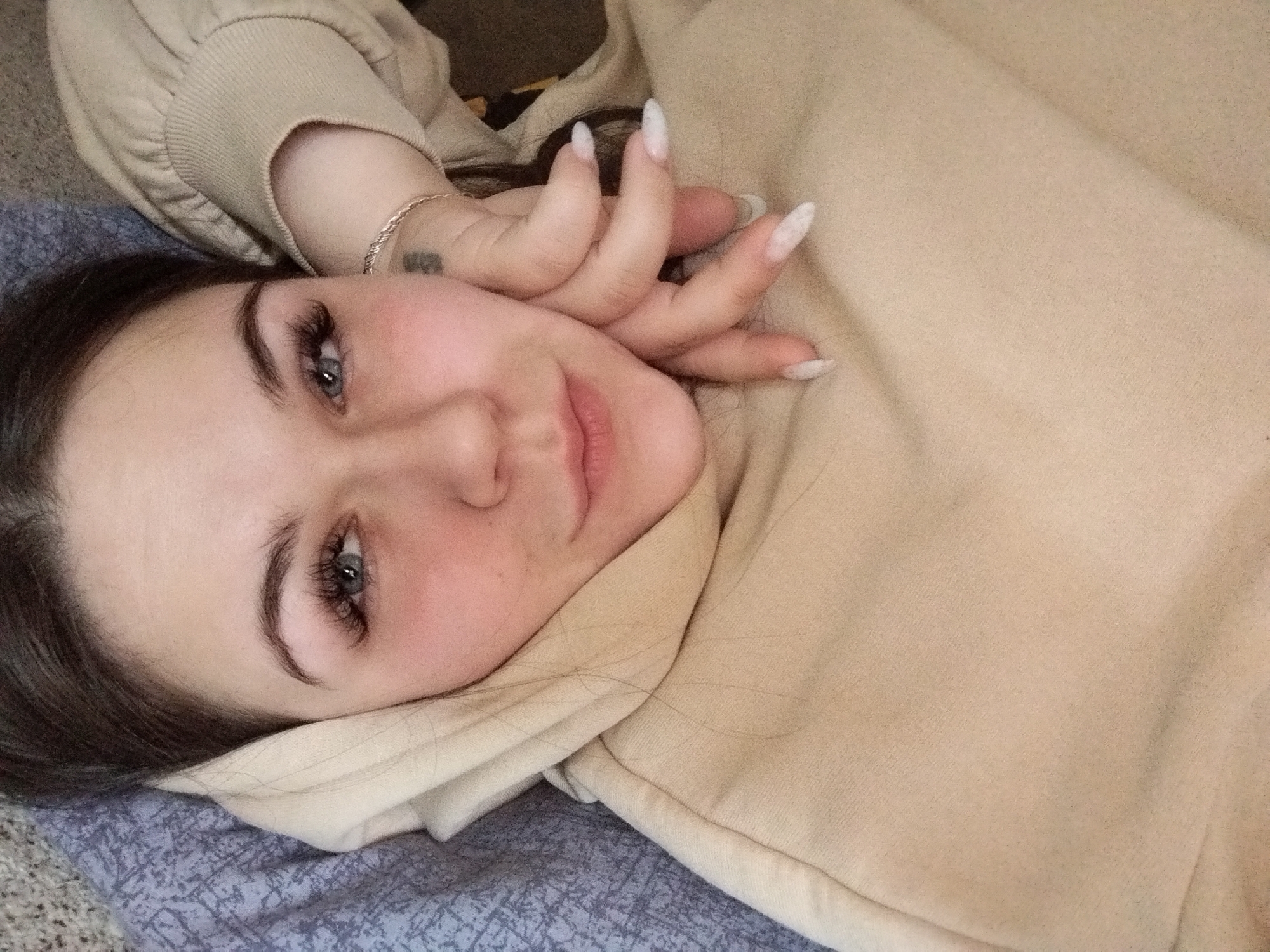 A woman with long dark hair and light skin lies on her back on a bed covered with a blue and gray patterned blanket. She is wearing a beige sweatshirt or hoodie, the hood of which is partially under her head. Her left hand, adorned with long, pointed nails, is near her face. A small tattoo is visible on her wrist. Her eyes are open, and she has long eyelashes. The location is unidentified, and there is no indication that this image was taken in Chaikovsky, Russia.