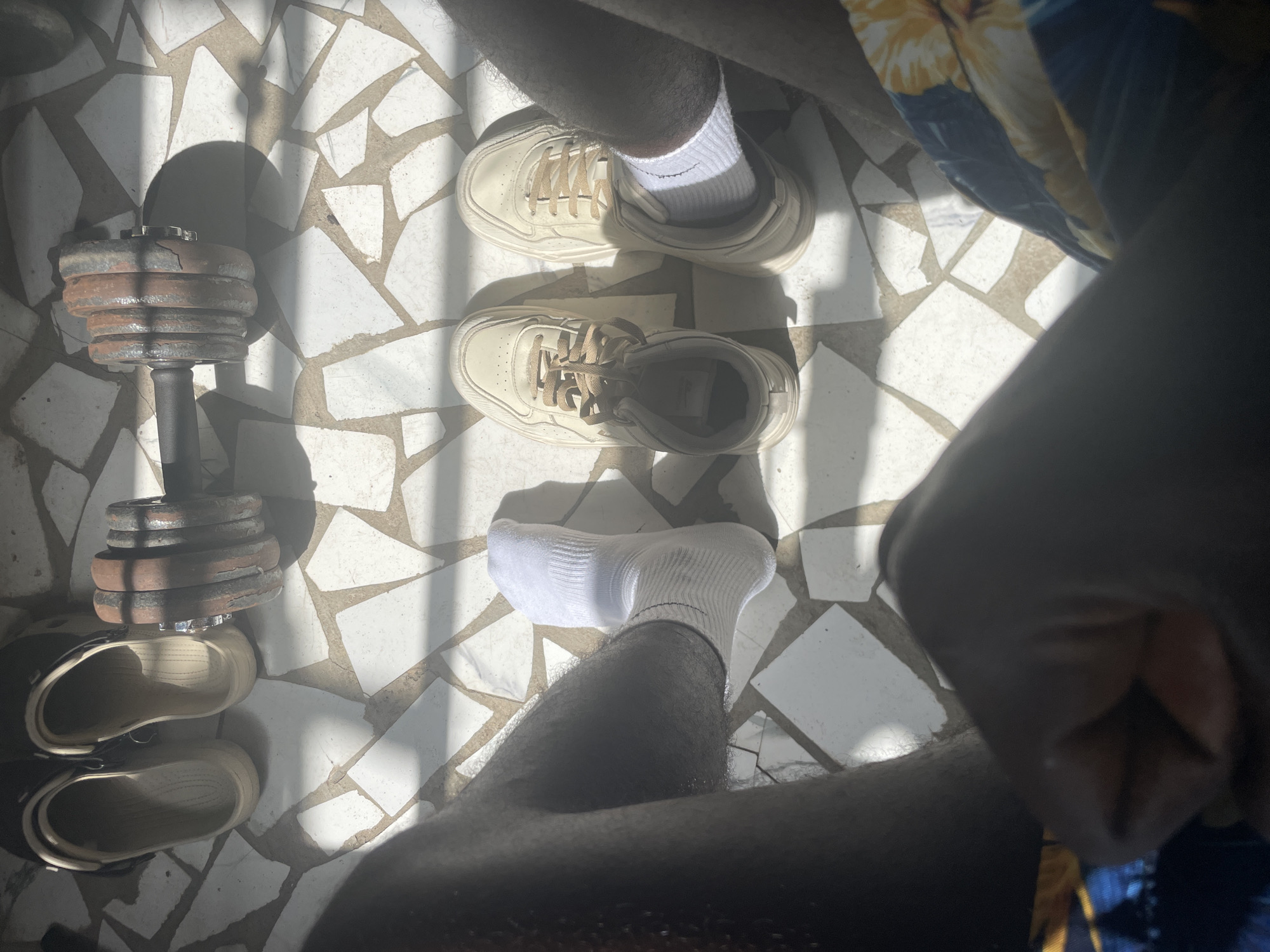The image, captured from a high angle looking down, presents a view of a person's lower body and surrounding objects on a distinctive tiled floor. The floor is composed of irregularly shaped white and dark grey/black tesserae, illuminated by strong, direct sunlight that casts sharp shadows, indicating a sunny day, likely morning or late afternoon, in Badagry, Nigeria.
The visible subject is a person with dark skin and hairy legs. One leg is extended, wearing a white ankle sock. Near this leg, a pair of cream or off-white sneakers with light brown laces are placed on the tiles. To the left of the frame, a heavy dumbbell with multiple rusty, circular weight plates rests on the floor. Below the dumbbell, a pair of beige, slip-on clogs or slippers is also present. On the right side, a portion of patterned clothing, possibly shorts, with a blue background and yellow floral designs, is visible. A blurred dark arm or hand is also seen on the far right. No specific activity is being performed, and no visible text is present.