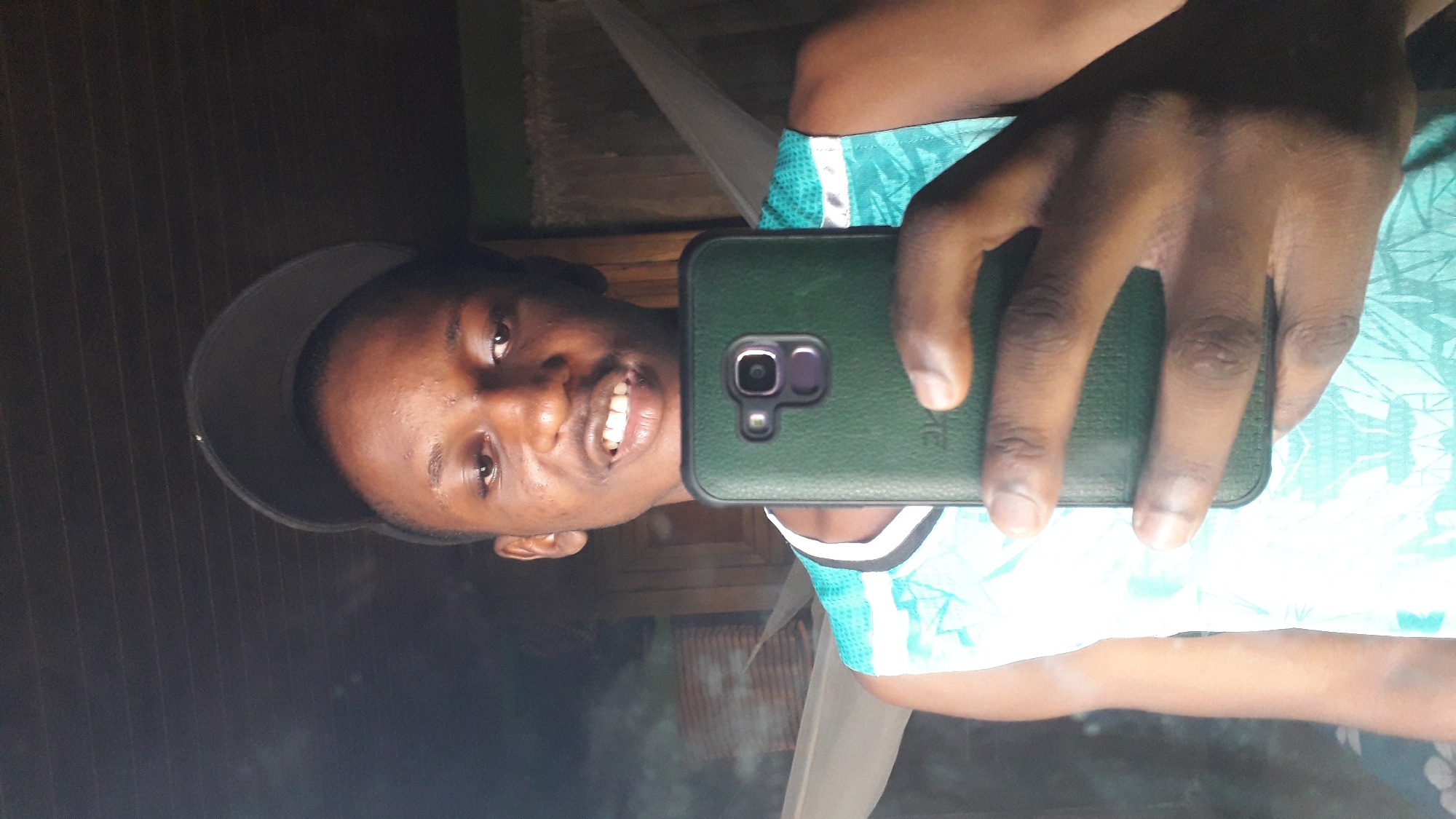 A person wearing a black baseball cap is holding a green-cased smartphone in their right hand, taking a selfie. The person is wearing a white and teal athletic shirt. They are smiling and facing the camera. The background appears to be a dimly lit interior space, potentially in Nkpor, Nigeria. A white mosquito net is partially visible.