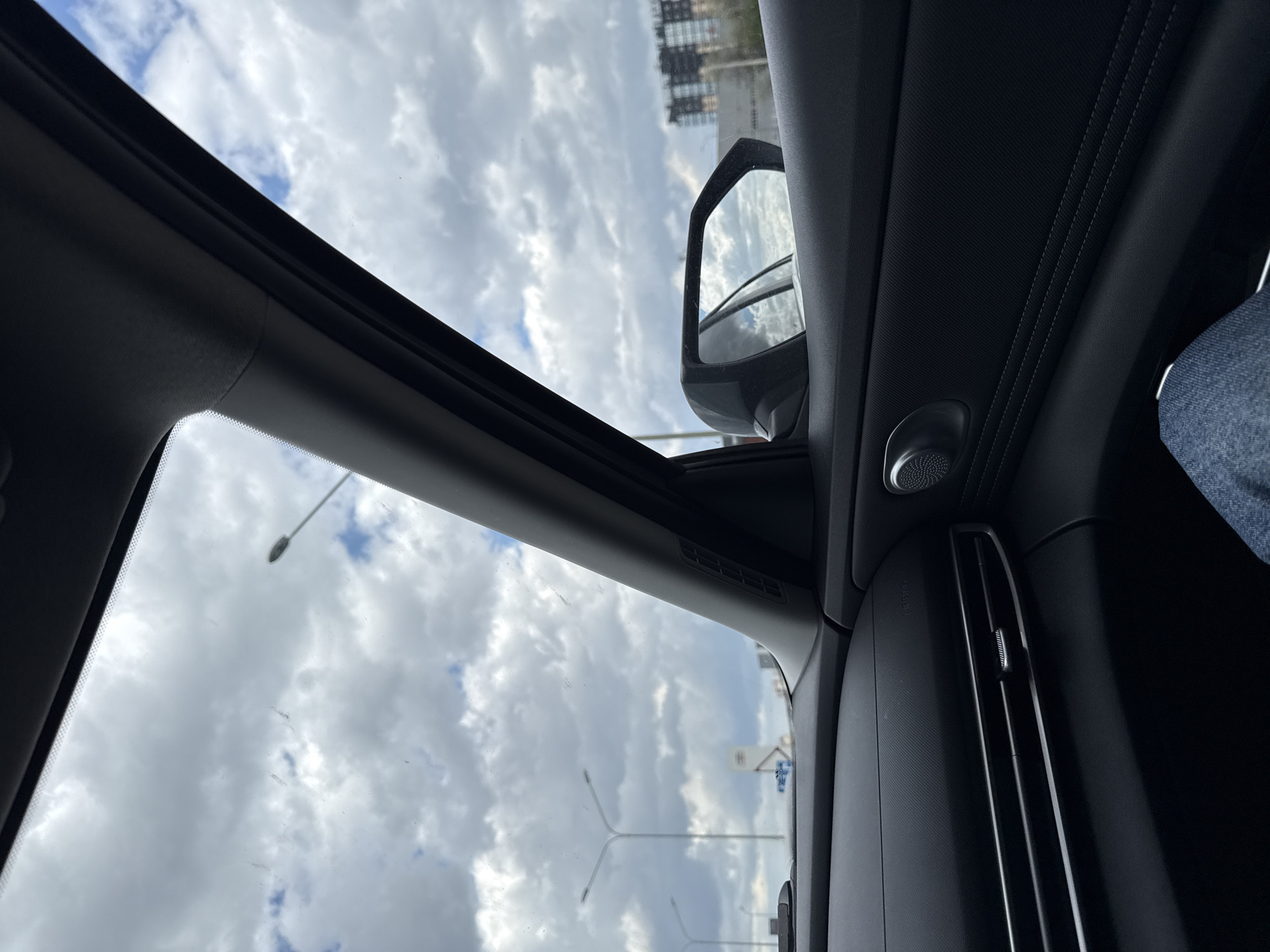 The image shows a view from inside a car, looking upwards and slightly to the left. The car has a sunroof, and through it, a cloudy sky is visible, along with street lights and building. A side mirror is visible, reflecting the sky and buildings. The interior of the car is dark, with a speaker and a ventilation system. A leg is visible. The location is unknown.