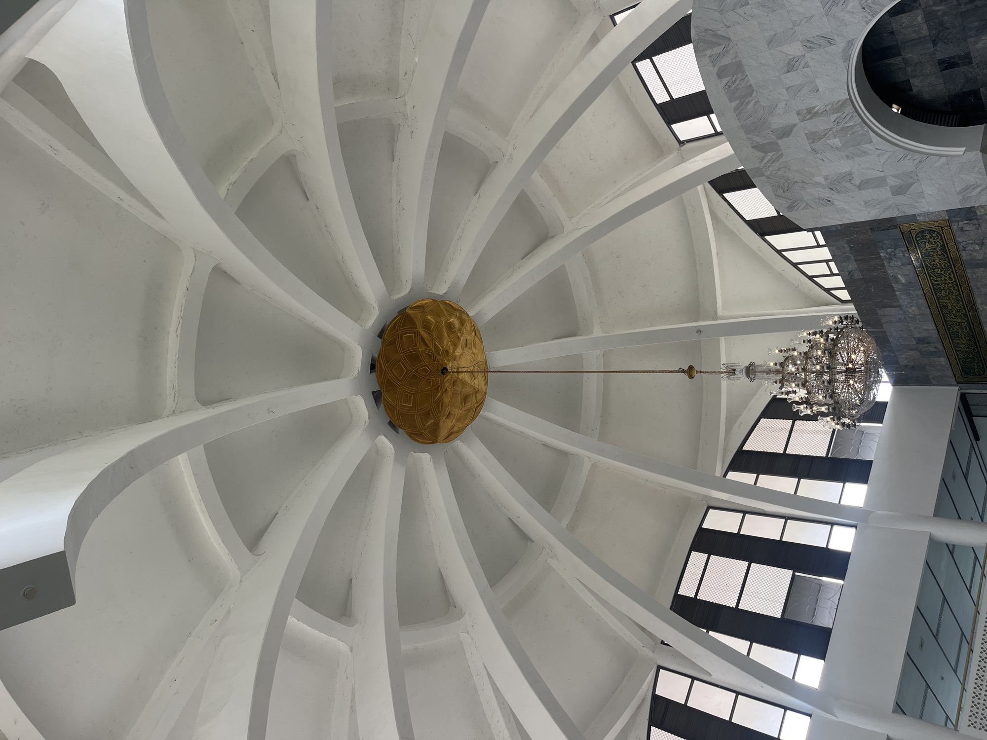 The image shows the interior of the Islamic Center mosque in Lhokseumawe, Indonesia, looking up towards the dome. The dome is white and composed of radial arches and panels. A large golden-colored ornamental sphere hangs suspended from the center of the dome. A chandelier hangs from the ceiling toward the right. There are windows along the sides of the interior. A section of marble or tiled wall is visible in the upper right corner. The shot is taken from a low angle.