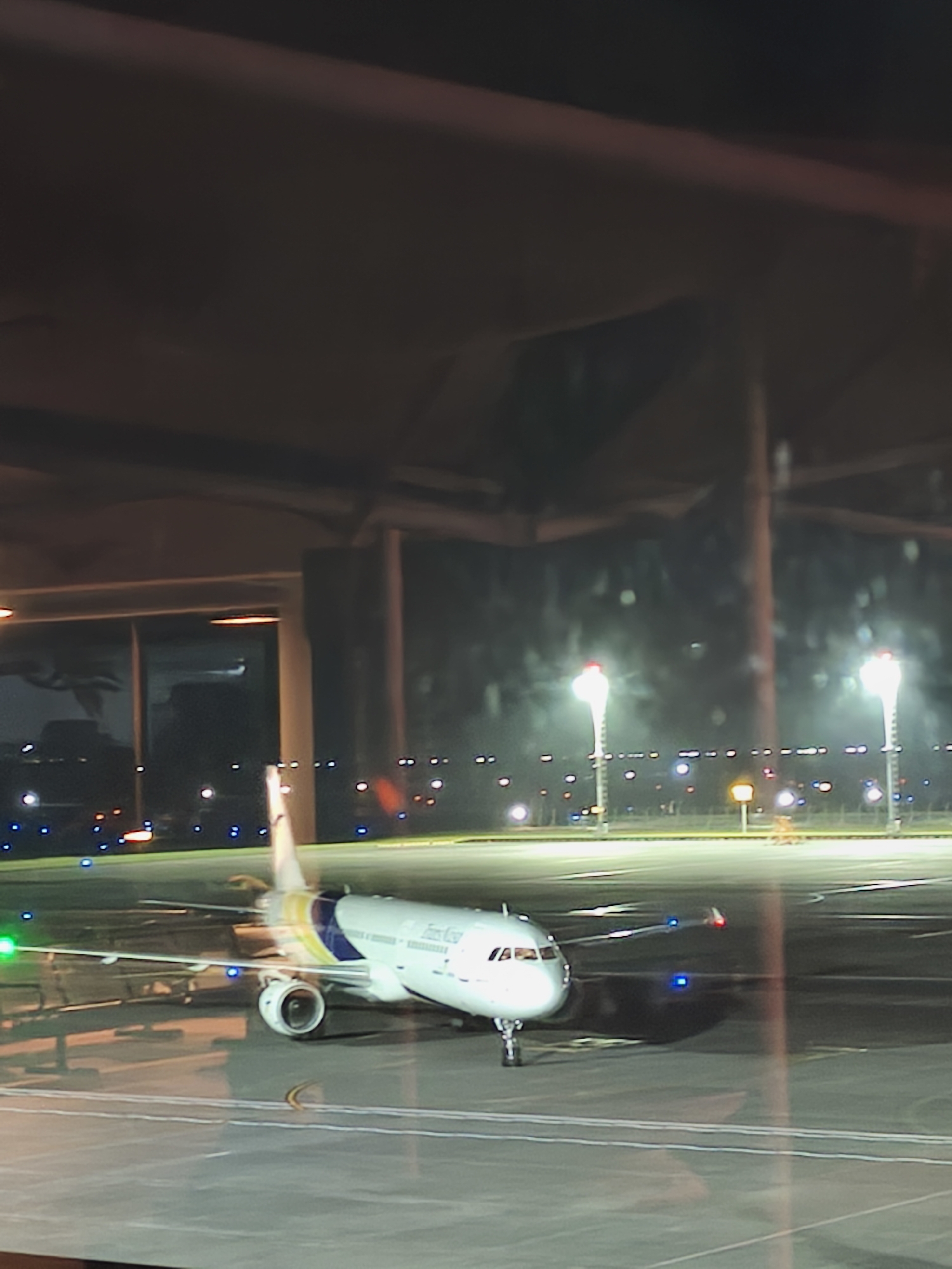 This media file shows an airplane on an airport tarmac at night. The airplane is white with blue and yellow accents on its tail. The word "Fly" is visible on the tail in white text. The scene is well-lit by airport lights, including runway lights and taller navigational lights, creating a bright and clear view despite the darkness. The airplane is parked on the tarmac, suggesting it is either preparing for departure or has just arrived. The overall setting is an airport environment, characterized by the presence of the airplane, tarmac, and lighting infrastructure. The time of day is clearly night. There are no people visible in the frame. The weather appears clear as the lights are sharp and not diffused by fog or rain. The location context of Kuta, Indonesia, is provided but not explicitly visible in the image itself.