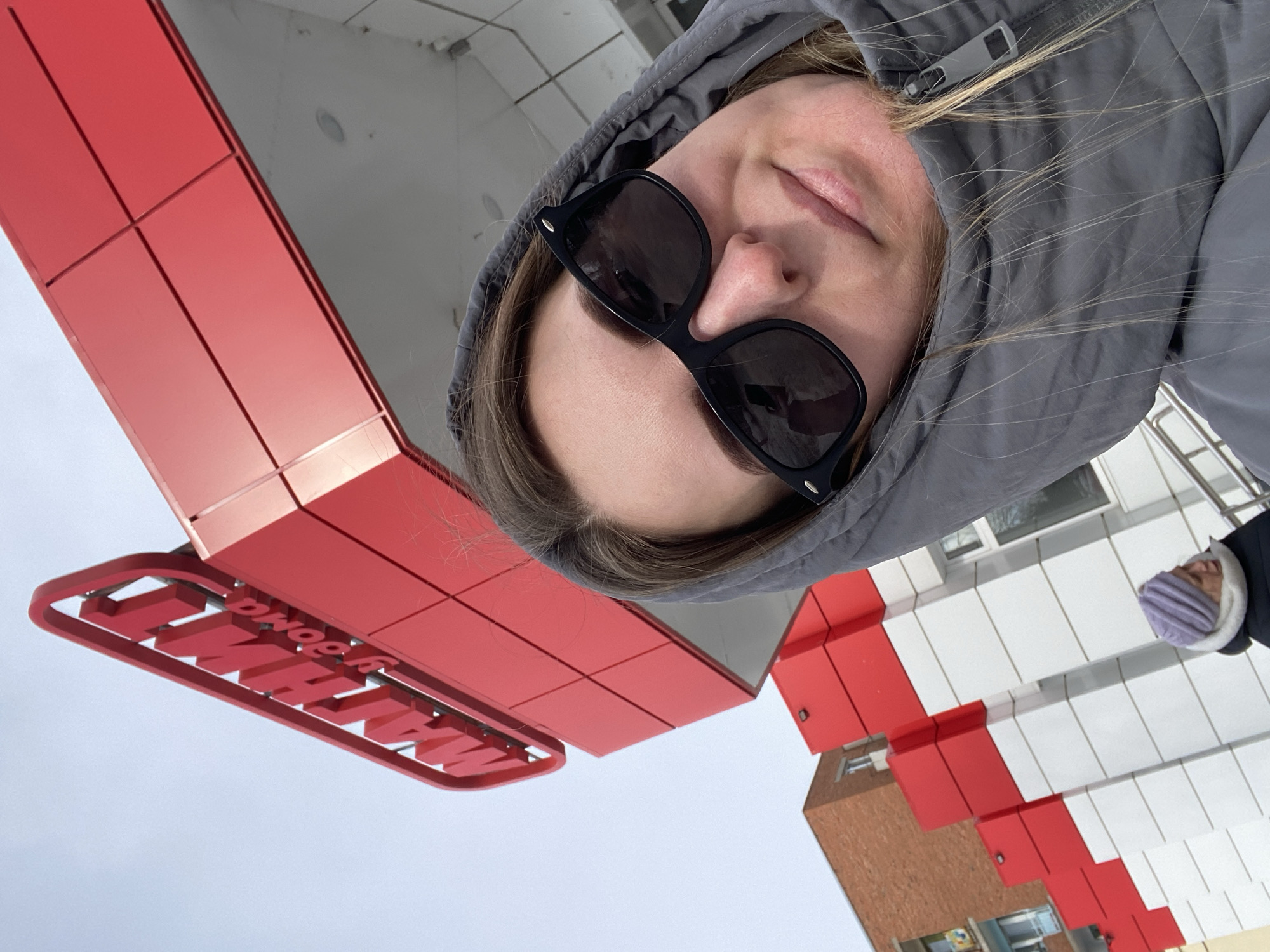 The image presents a close-up, low-angle outdoor scene in Noviy, Russia. The primary subject, a female, occupies the lower right portion of the frame, wearing black Wayfarer-style sunglasses and a grey hooded puffer jacket, with light brown hair visible around her face. In the upper left background, a prominent red sign with three-dimensional Cyrillic letters reads "МАГНИТ у дома" (Magnit at home), identifying a supermarket. The building's facade beneath the sign features a modern design of alternating red and white panels. A secondary individual is partially visible in the lower-left background, wearing a light purple knit hat and a dark jacket, facing away from the camera. Further in the background, a multi-story brick residential building is observable. The sky is light and overcast, indicating an urban environment under daylight conditions, suggesting cool weather.