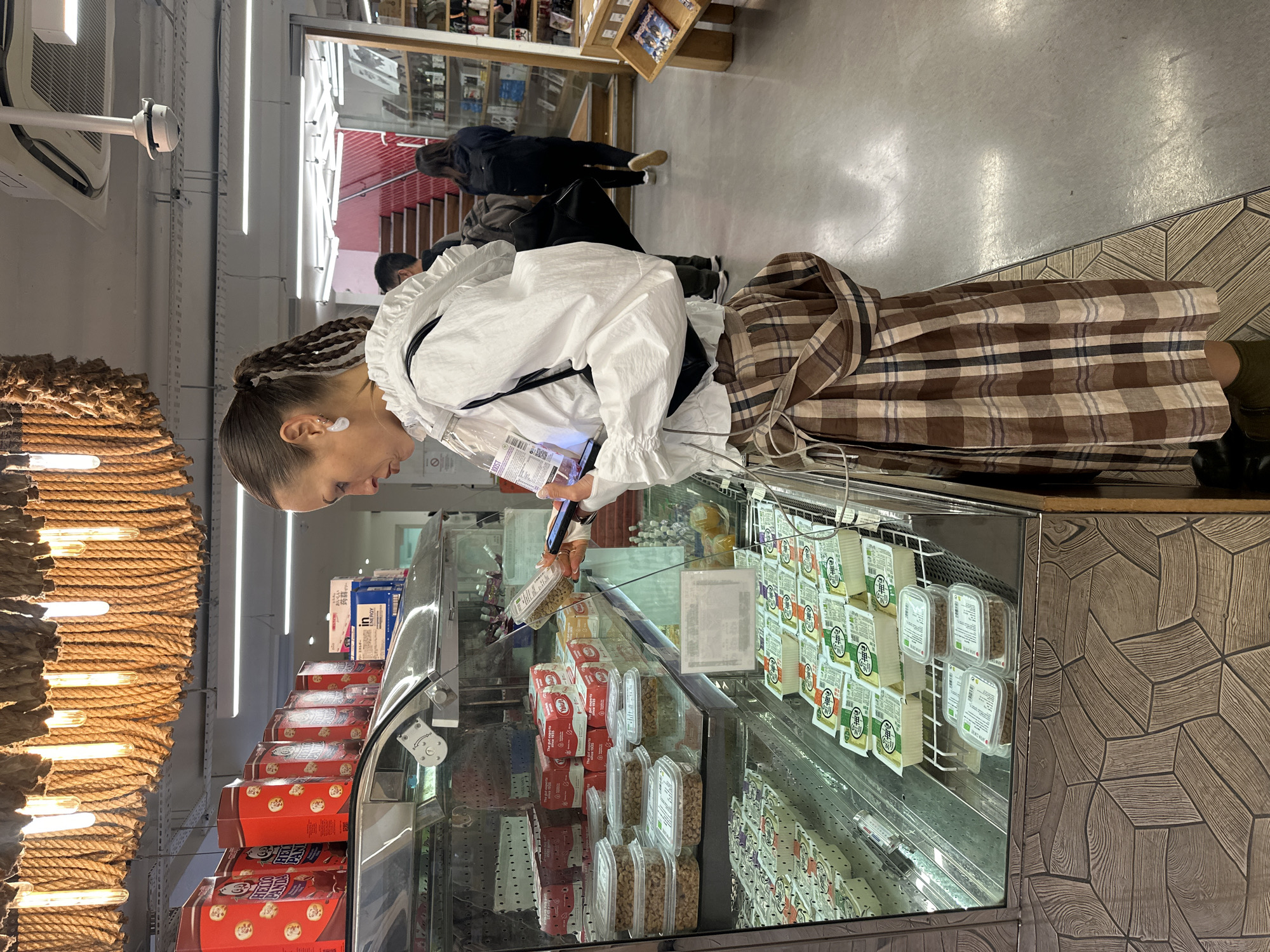 Here is a detailed description of the image:
**Scene Overview:**
The image captures an indoor scene within what appears to be a modern grocery store. The focus is on a woman browsing a refrigerated display case filled with various food products. There are other people walking by in the background.
**People:**
* **Woman:** The main subject is a woman standing in front of the display case. She is wearing a distinctive outfit consisting of a white ruffled jacket or blouse and a brown plaid skirt. She has her hair styled in braids and is wearing what appears to be an earpiece. She is holding a container in one hand and a smartphone in the other, seemingly reading or comparing information about the product.
* **Other People:** In the background, there are other people walking. One person is wearing a black coat.
**Objects:**
* **Refrigerated Display Case:** The display case is filled with a variety of packaged food items, including what looks like nuts, tofu, and other snacks or ingredients. The labels of the products are not fully legible, but some seem to have Japanese writing on them. Boxes of The Gut Expert's Yakult snacks are visible in the refrigerated display.
* **Rope Display:** To the left of the woman, there is a decorative display made of thick twisted rope with embedded light fixtures.
* **Lighting:** The store is well-lit with long linear fluorescent lights.
* **Smartphone:** The woman is holding a black smartphone while examining a package.
* **Grocery Store Shelves:** In the background, you can see more shelves with products.
**Actions:**
* **Browsing/Comparing:** The woman is actively examining products in the display case, likely comparing information or making a purchasing decision.
* **Walking:** Other people are walking around in the background, presumably shopping.
**Environment:**
* **Grocery Store:** The setting is a modern grocery store, characterized by clean lines, organized displays, and bright lighting.
* **Flooring:** The floor near the refrigerated display has a patterned, geometric design.
The image is taken from a side angle, providing a clear view of the woman and the display case, giving a sense of a normal, everyday shopping scene within the grocery store.