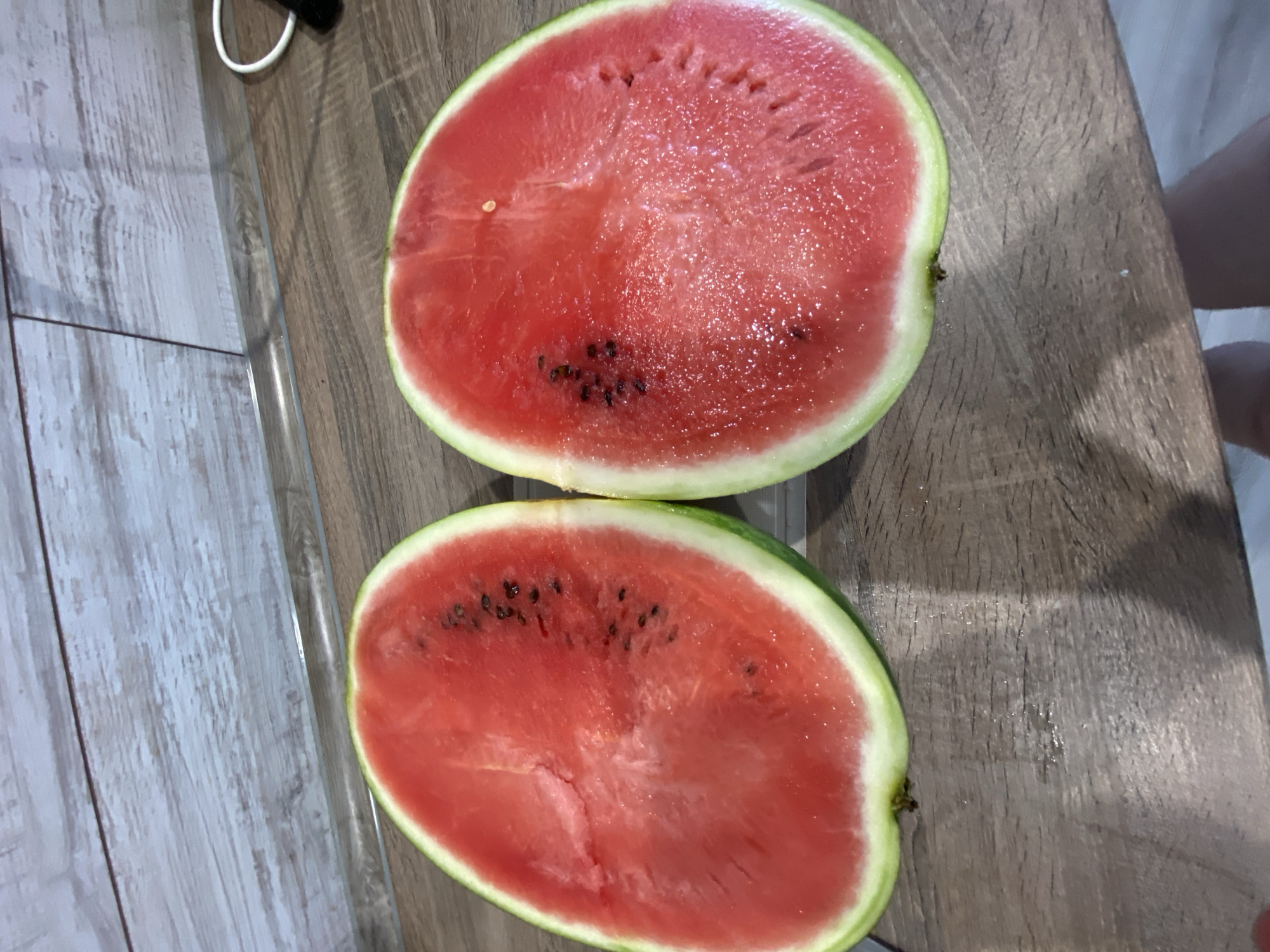 The image shows two halves of a watermelon, cut lengthwise, resting on a wooden surface. The watermelons are bright red with visible seeds and a green rind. The wooden surface appears to be a cutting board or table. In the background, a white cord is visible, possibly connected to an electronic device. The lighting suggests it is daytime, and the overall setting seems to be indoors, likely a kitchen or dining area. There are no people clearly visible in the image, though a hint of a person's leg or arm is present in the upper right corner. The scene is simple and focuses on the fruit. There is no visible text in the image.
