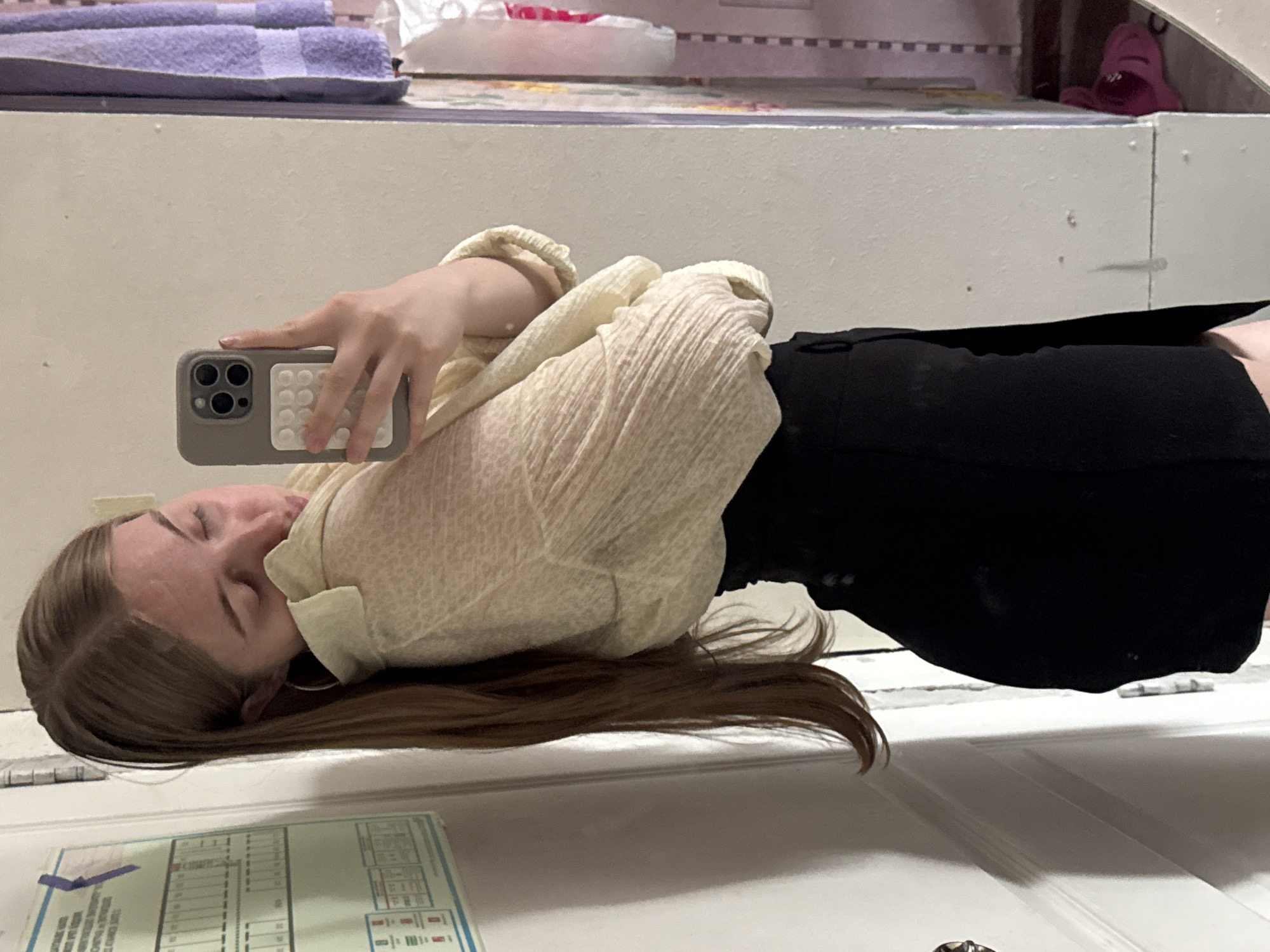 A young woman with long, brown hair is standing in a room, taking a selfie in a mirror. She is wearing a light-colored, textured blouse and black high-waisted pants. Her head is tilted down, and she is looking at her phone, which is held in her right hand. The phone has a grey case with a pop socket on the back. Behind her, on the left, is a door with some informational posters attached. To her right, there is a mirror reflecting her and a glimpse of a purple towel and other items in the background. The environment appears to be a domestic setting, possibly a bedroom or hallway, and is consistent with a location in the city of Perm, Russia, based on the presence of the informational posters which are in Cyrillic script and similar to those found in Russian apartment buildings.