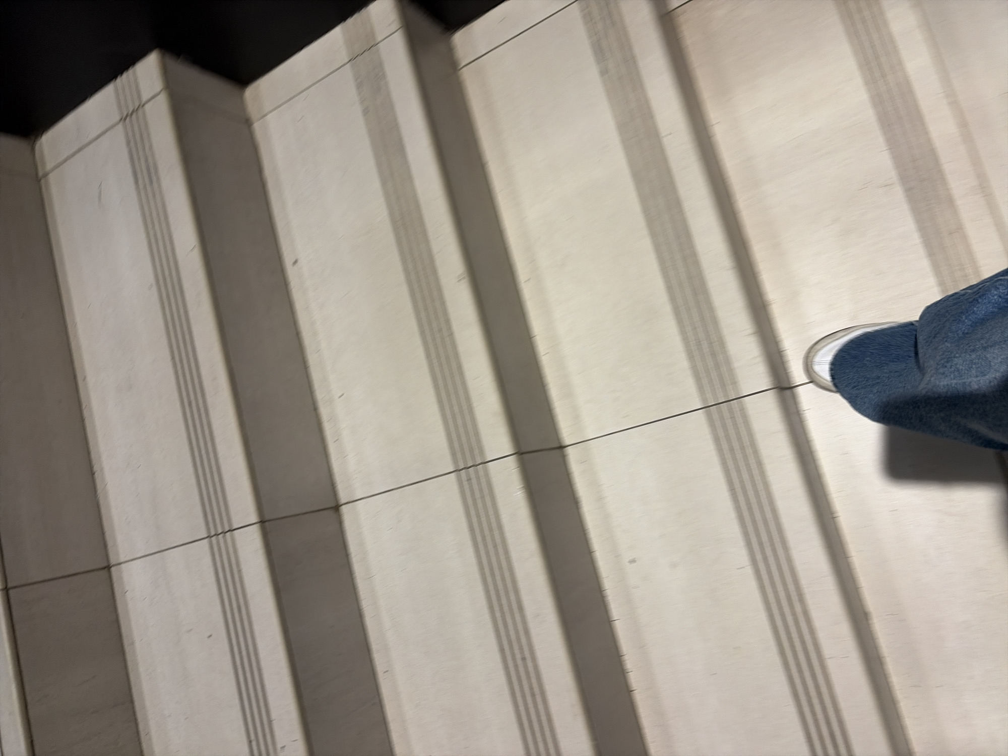 The image shows a person's foot and lower leg, clad in blue jeans and a white sneaker, stepping onto a light-colored tiled staircase. The staircase has a non-slip textured strip on each step, running horizontally across its width. The steps are clean and appear to be made of ceramic or stone tiles. The lighting suggests an indoor environment, possibly a public building or a modern home. There are no specific visual cues in the image that would allow for the determination of its location within the city of Moscow, Russia.