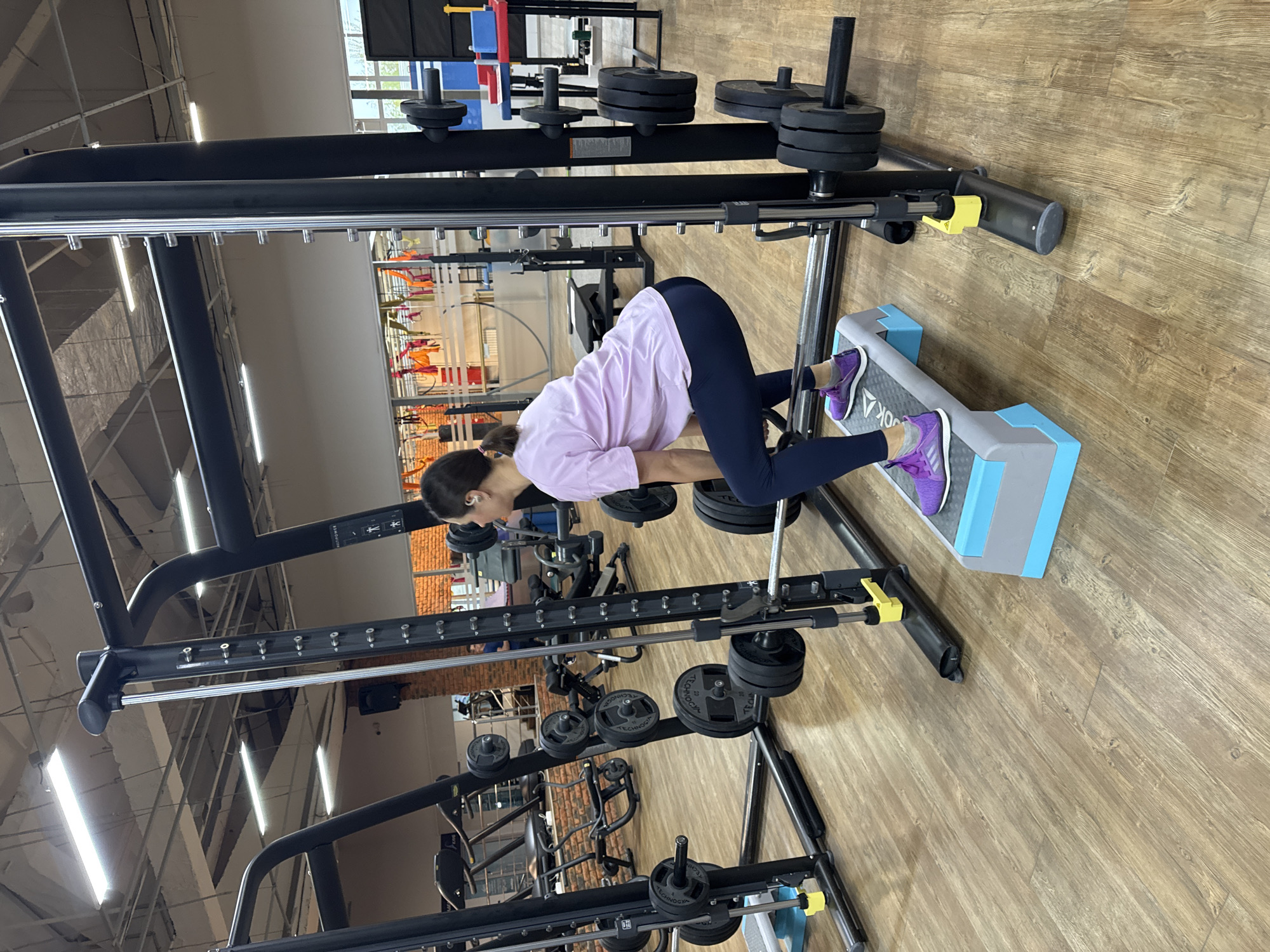 A woman is performing a weightlifting exercise at a gym. The location is likely in Chaikovsky, Russia. She is standing on a Reebok step platform, leaning forward while holding a barbell on her back. She is wearing purple sneakers, dark leggings, and a light pink t-shirt. The gym contains various weightlifting machines with weights. The floor is made of wood.