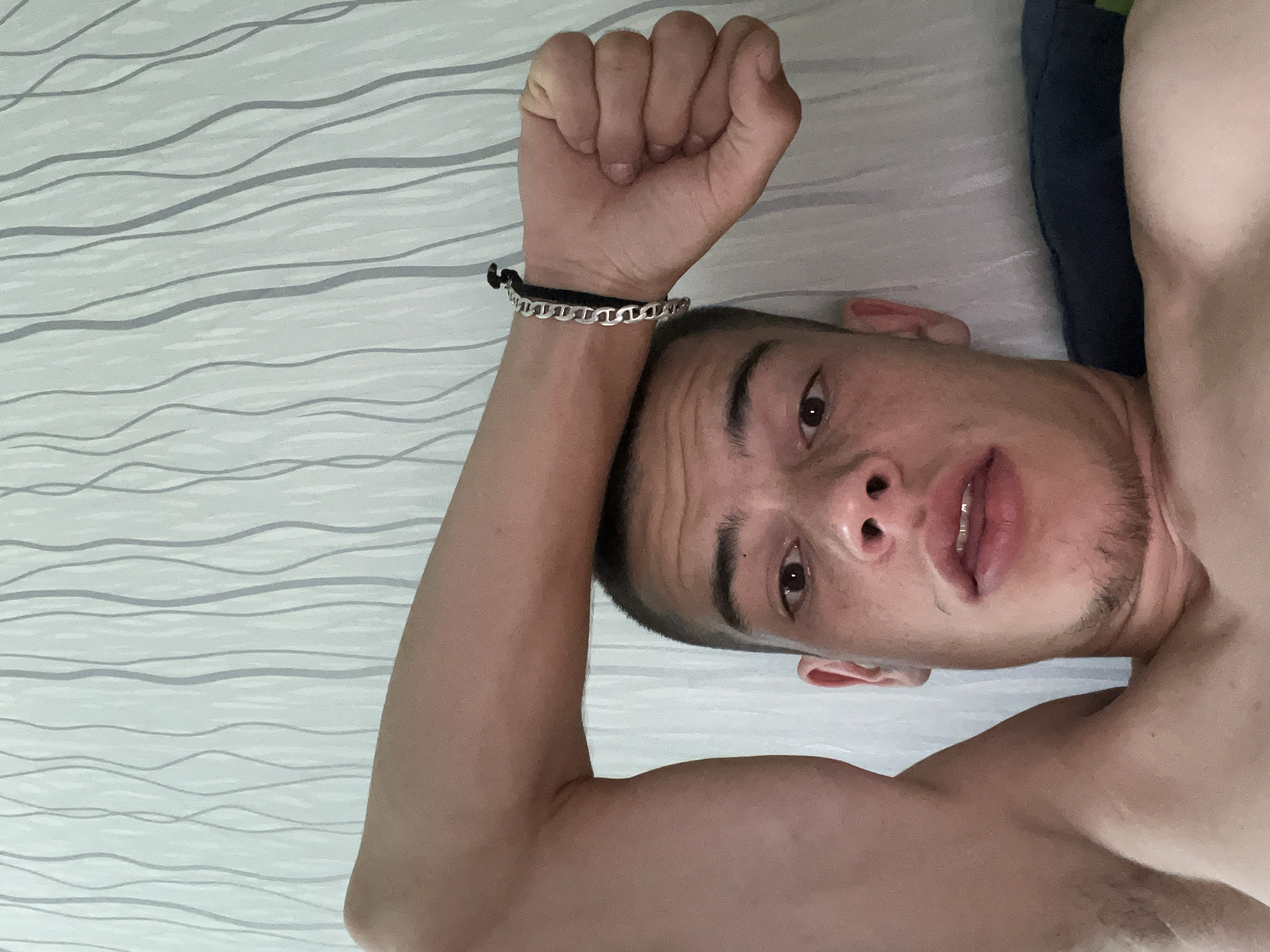 The image is a close-up, low-angle selfie of a young man lying down. He has short, dark hair and a light beard. His right arm is raised and bent, with his forearm resting on his forehead. He is wearing a silver chain bracelet on his wrist. His mouth is slightly open, and he appears to be looking at the camera. The background is a wall with a light-colored wallpaper featuring a pattern of thin, grey, wavy vertical lines. The scene is indoors, with a dark blue pillow visible at the bottom right of the frame. There is no indication of the specific location within the city of Sopot, Bulgaria.