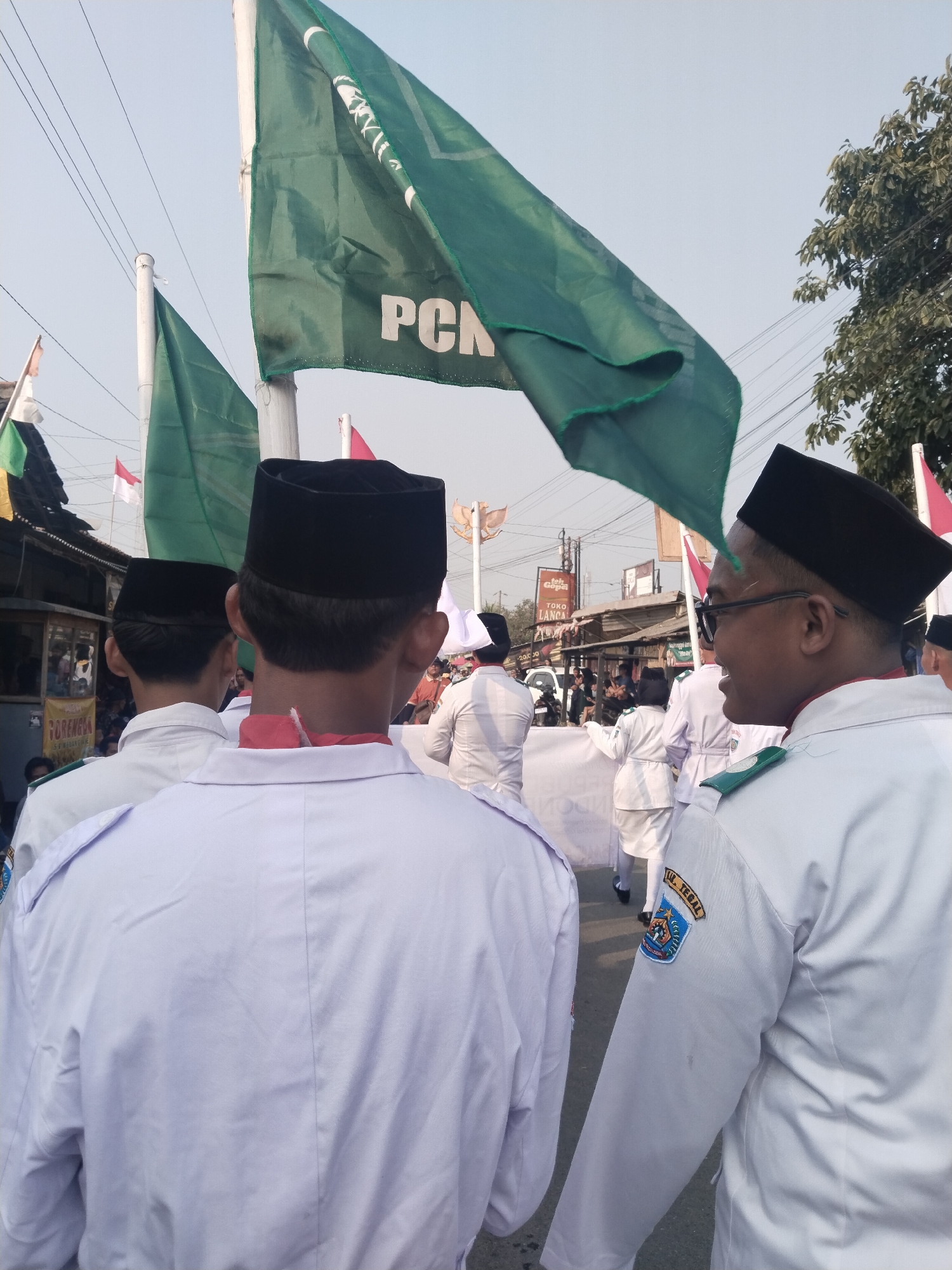 In Margasari, Indonesia, a group of individuals wearing white shirts and black caps participate in an outdoor event, possibly a parade or procession. Dominant in the frame is a large green flag with white lettering that appears to say "PcN". Other green flags are also visible. Several people are seen from behind, also wearing white uniforms and black caps. One individual in the foreground on the right is wearing glasses and smiling. Another individual on the left also wears a black cap and a red scarf. In the background, there are buildings, trees, and a clear sky. The scene suggests a community gathering or a celebration.