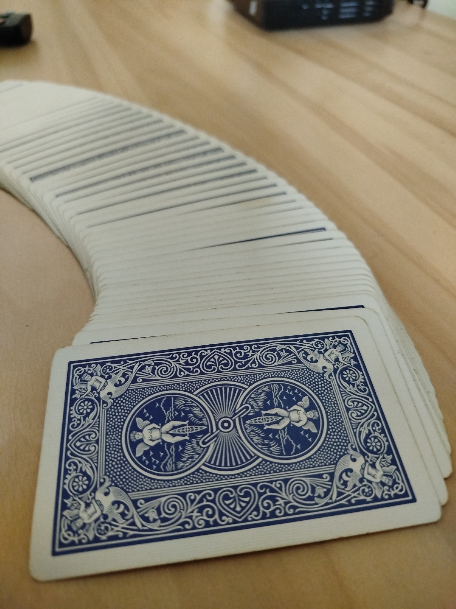 A deck of playing cards is fanned out on a wooden surface. The back of the bottom card is visible, featuring a blue design with cherubs riding bicycles. The location is unknown.