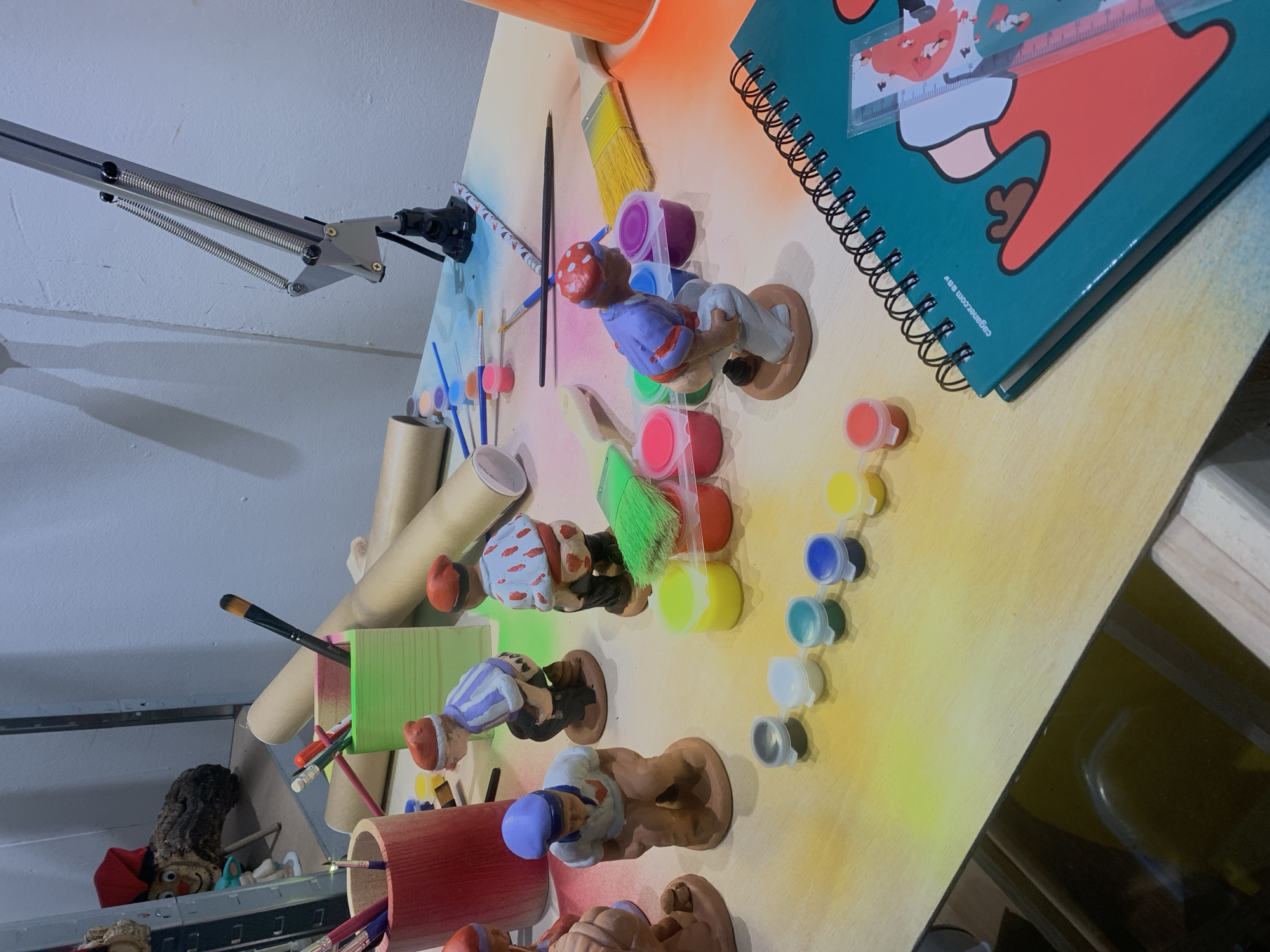 The image depicts a tabletop scene with various art supplies and painted gnome figurines. There are pots of paint, brushes, a notebook, and containers holding art tools. The gnomes appear to be in different stages of painting. The presence of a notebook with a Catalan-language website printed on it, "caganer.com eav", suggests the location is in Barcelona, Spain, as Caganer figurines are a traditional Catalan Christmas decoration. The items are arranged on a light-colored surface, possibly a table or desk, with a flexible arm lamp extending from the upper-left corner. A shelf with a doll is visible in the background.