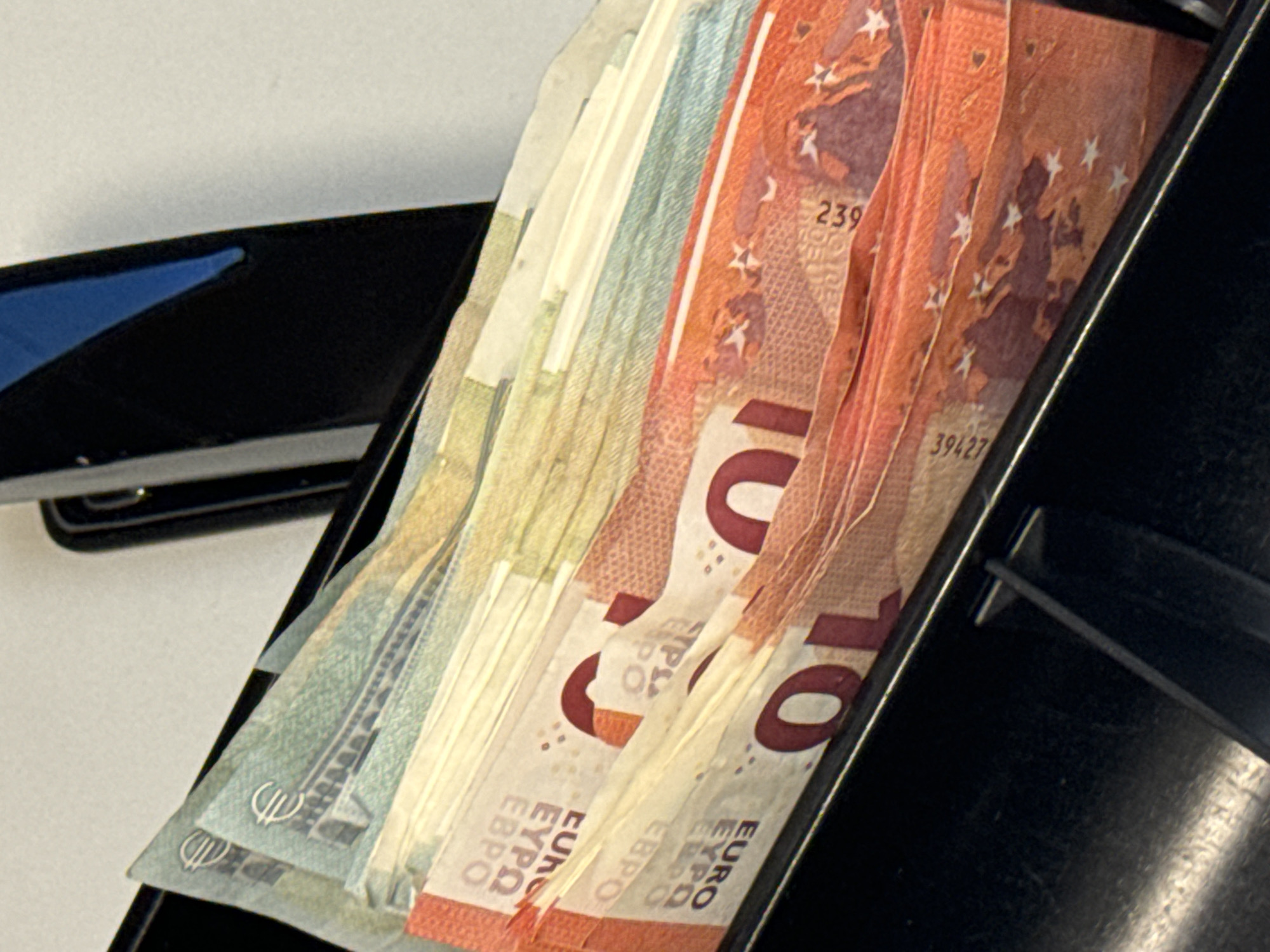 The image shows a stack of Euro banknotes, primarily 10 Euro notes, partially inserted into a black cash register drawer. A stapler is visible to the left of the drawer. The location is unknown.