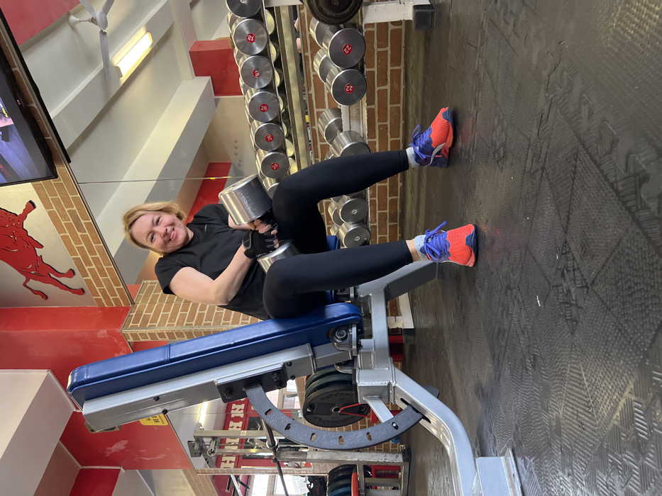 An interior image taken in a fitness center located in Saint Petersburg, Russia, depicts a Caucasian woman with blonde hair, smiling, and seated on a grey and blue adjustable weight bench. She is wearing a black short-sleeved t-shirt, black leggings, black fingerless workout gloves, white ankle socks, and vibrant athletic shoes with orange/red and blue/purple coloring. Her right hand, gloved, firmly grips a large, polished chrome dumbbell, with her forearm resting on her right thigh.
The background features a wall composed of red-painted sections interspersed with reddish-brown brick patterns. A prominent red silhouette of a charging bull is affixed to a red wall section above the brickwork. To the right of the woman, a two-tier metal rack holds a series of chrome dumbbells, with red numerical labels visible on their ends (e.g., 22, 26, 28, 30). A flat-screen television is mounted high on the wall. The white ceiling displays rectangular fluorescent light fixtures and a white ceiling fan. The floor is covered with black, textured rubber gym mats. On the left side of the frame, a metal rack holding barbell weight plates is partially visible, and large wall-mounted mirrors reflect portions of the gym environment, including additional equipment and red lettering.