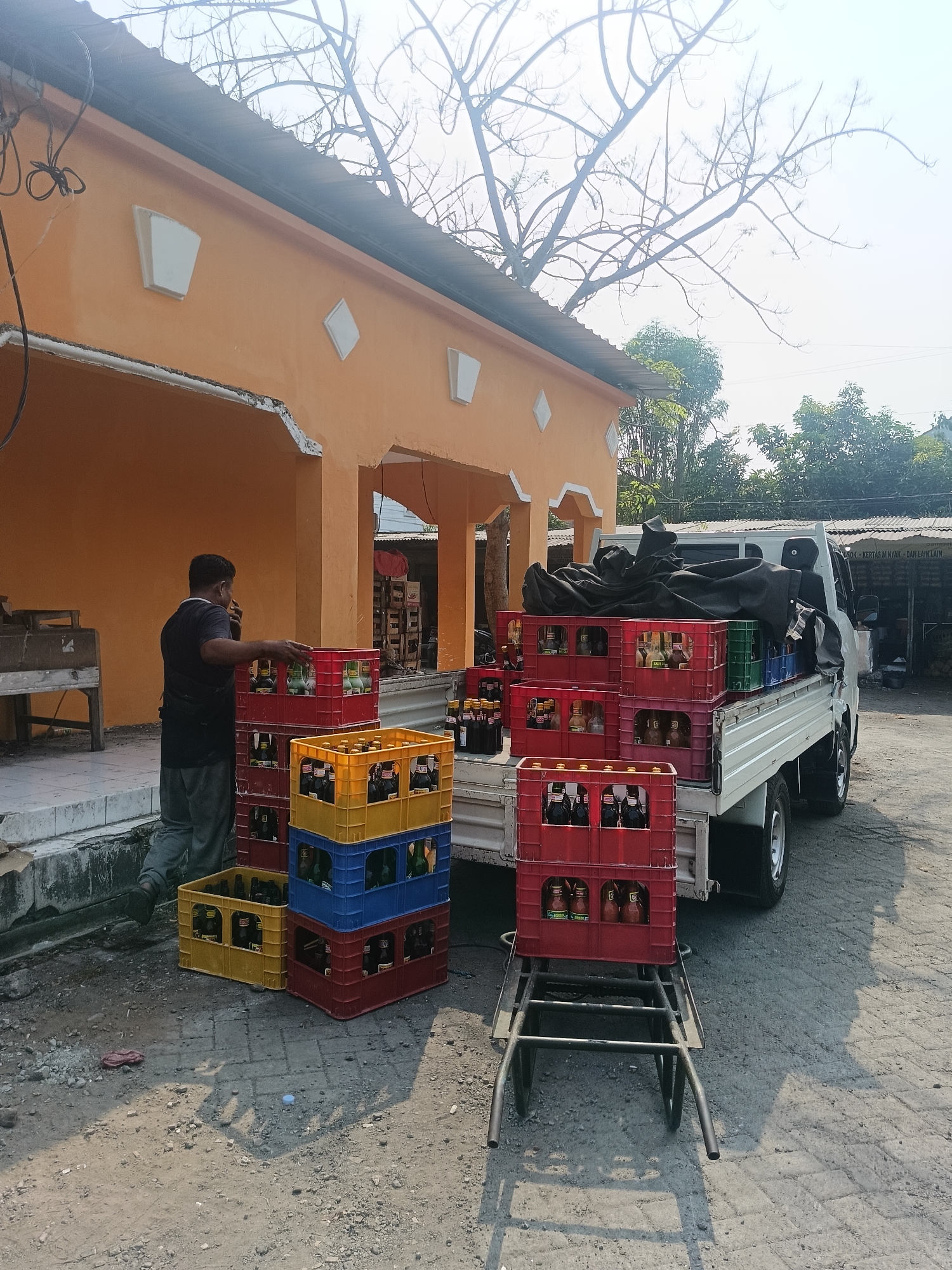 An orange building with a corrugated metal roof stands in Ketajen, Indonesia. In front of the building, a man in a dark t-shirt and grey pants is on his phone while standing next to stacks of plastic crates filled with glass bottles. Some crates are red, some are yellow, and one is blue. A white utility truck is parked nearby, loaded with more crates and a black tarp. The ground is covered in gravel and some paving stones. The scene appears to be a commercial area, possibly a distribution point for beverages.