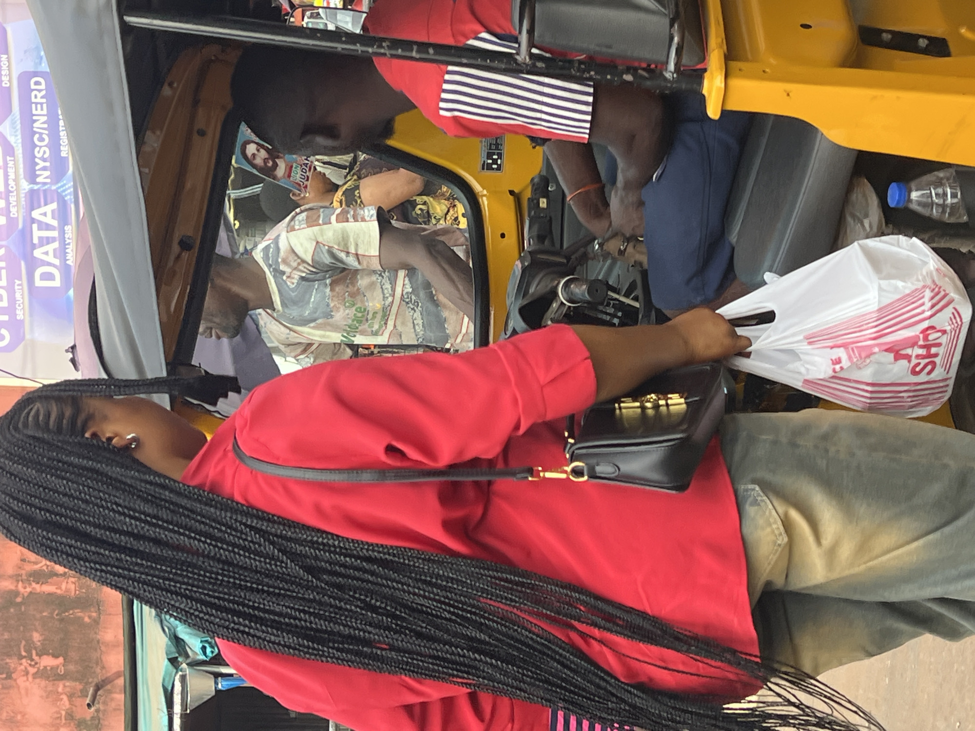 The image shows a group of people inside and around a yellow three-wheeled auto-rickshaw, commonly known as a Keke Napep, in Nkpor, Nigeria. The foreground features a woman with long, braided black hair, wearing a bright red long-sleeved top and greyish-green pants. She has a black handbag with gold hardware slung over her shoulder. Her left hand is visible, holding a white plastic shopping bag with red lettering.
Inside the Keke, a person wearing a red shirt with blue and white striped sleeves is visible in the background, leaning forward. Another person, in the center of the auto-rickshaw, is wearing a patterned shirt and appears to be looking towards the front. The dashboard and steering wheel of the Keke are also visible. To the right, a blue plastic bottle is placed on the floor.
A large banner with text in purple and white is partially visible on the left side of the image. Text visible includes "CYBER", "DATA", "DEVELOPMENT", "ANALYSIS", "NYSC/NERD", "DESIGN", and "REGISTE". The scene appears to be outdoors during daylight, though the weather is not discernible. The overall impression is of daily life and transportation in a Nigerian urban setting.