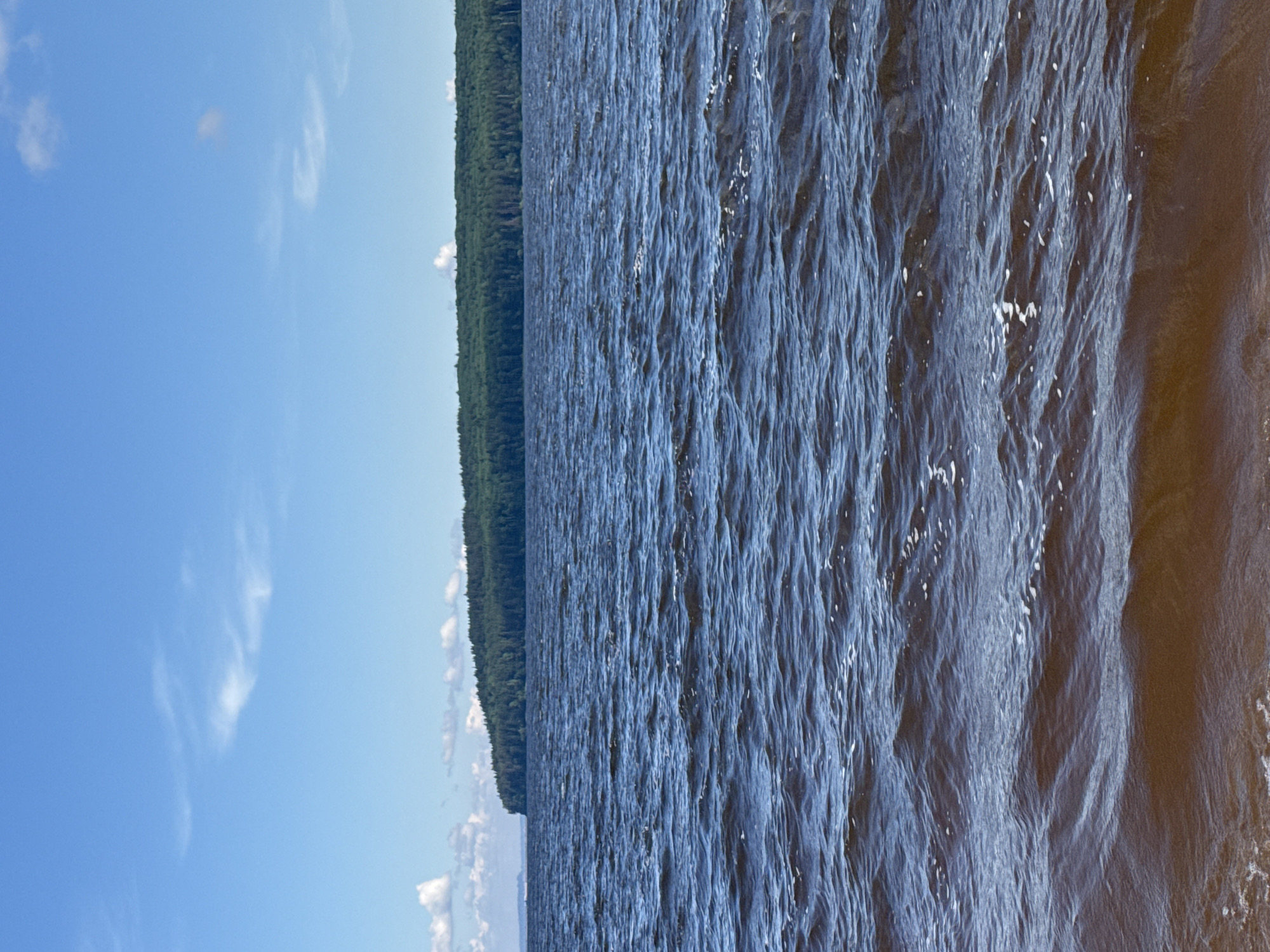 This media file presents a panoramic landscape view of a vast body of water, likely a lake or large river, under a bright blue sky. The image appears to be rotated, but if viewed with the sky at the top, it depicts a clear day.
In the upper portion of the frame, a wide expanse of vibrant blue sky stretches across, adorned with a few wispy, scattered white clouds, suggesting fair weather. Below the sky, a distant, dark green forest forms a distinct, continuous horizon line, indicating a dense tree line bordering the water.
The majority of the image is dominated by the water, which appears deep blue to almost dark grey in varying shades. The surface is heavily textured with numerous small, agitated waves and ripples, indicating a light to moderate breeze. Many small white flecks, consistent with foam or tiny whitecaps, are scattered across the water's surface, particularly in the mid-ground and foreground, adding to the sense of movement. The water closest to the viewer (bottom of the corrected image) exhibits more pronounced wave patterns and darker tones, possibly due to depth or light conditions.
There are no visible people, man-made objects, or discernible text within the scene. The setting suggests a natural, expansive environment, possibly a large lake or river in the vicinity of Chaikovsky, Russia, such as the Kama Reservoir. The lighting indicates it is daytime, likely mid-morning or afternoon, given the clear skies and apparent sun brightness. The overall mood conveyed is one of natural tranquility and vastness.