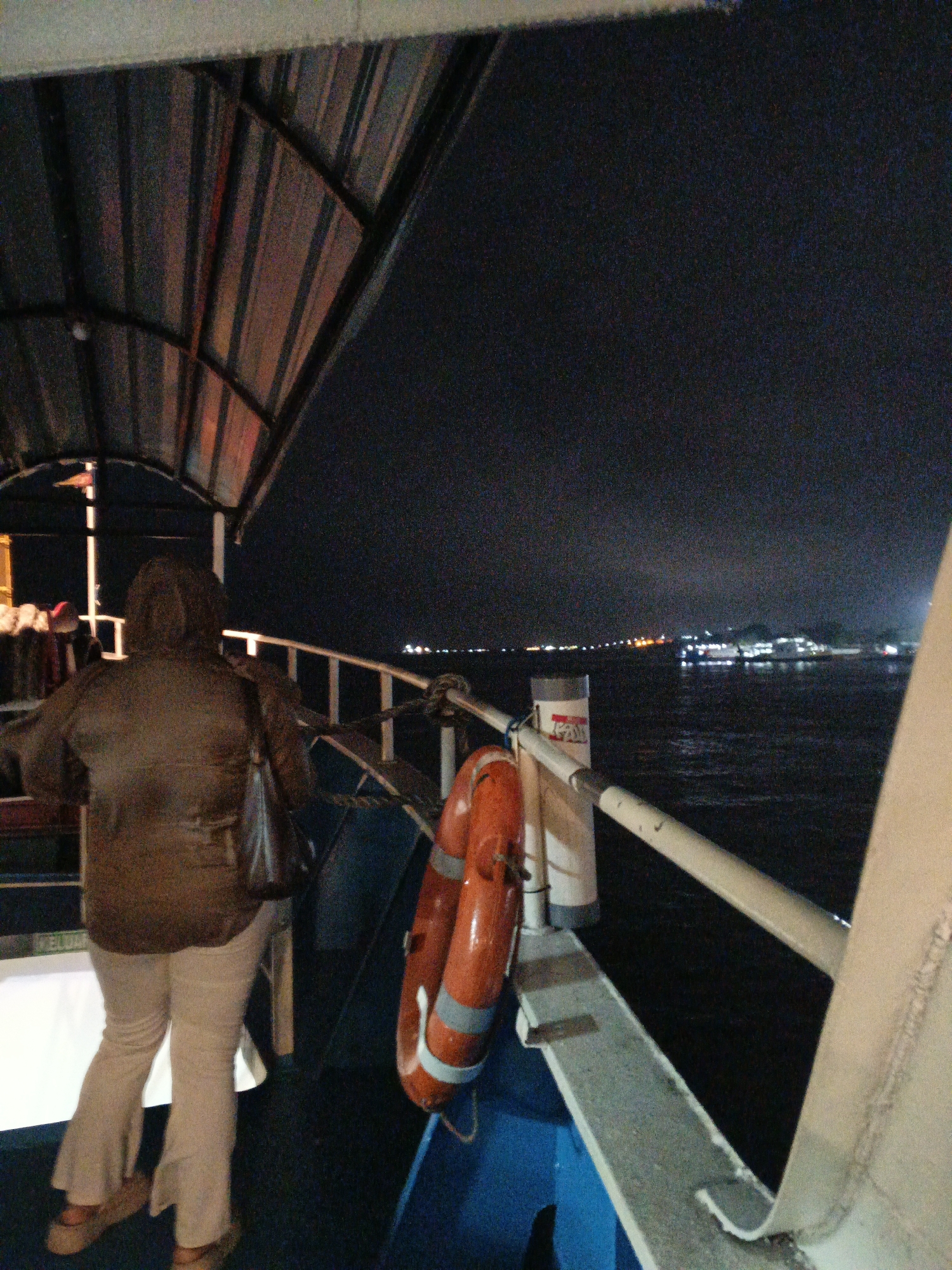 The image depicts a nighttime scene on a ferry, likely in the waters near Banyuwangi, Indonesia, given the illuminated coastline visible in the distance. The foreground features a person standing on the deck, viewed from behind. They are wearing a brown hooded jacket and light-colored flared pants, with a dark bag slung over their shoulder. To their right, a bright orange life preserver with light blue stripes is hanging on the railing. The ferry's deck is covered in a dark blue carpet. A metal roof structure with exposed beams and lighting is visible overhead. The water is dark, reflecting the distant lights of the city. The overall impression is one of being on a moving vessel at night, with the urban lights of Banyuwangi serving as a backdrop.